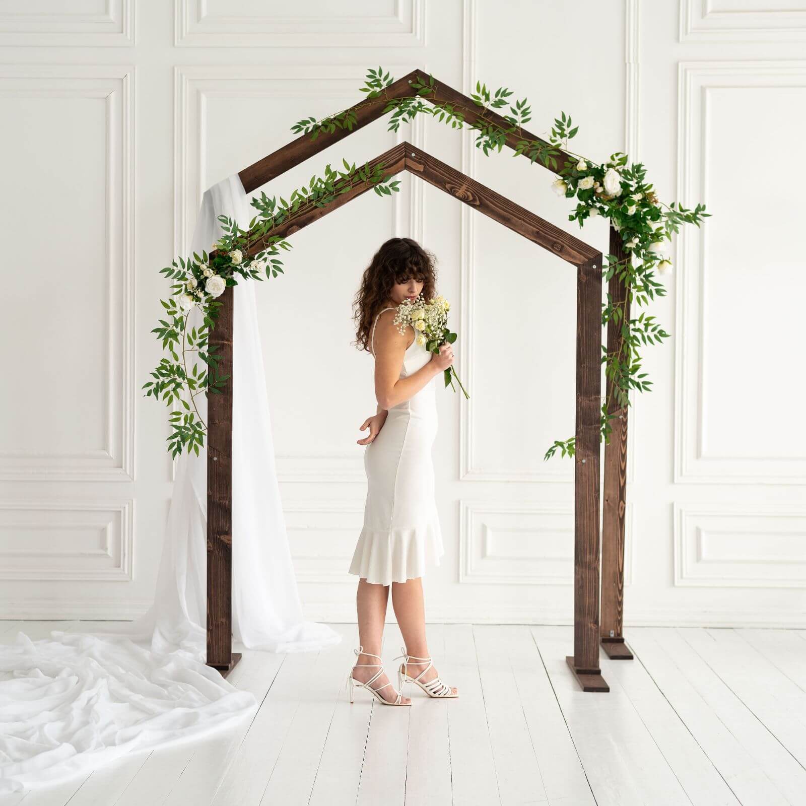 Bride standing under dark walnut pentagon arch decorated with greenery.