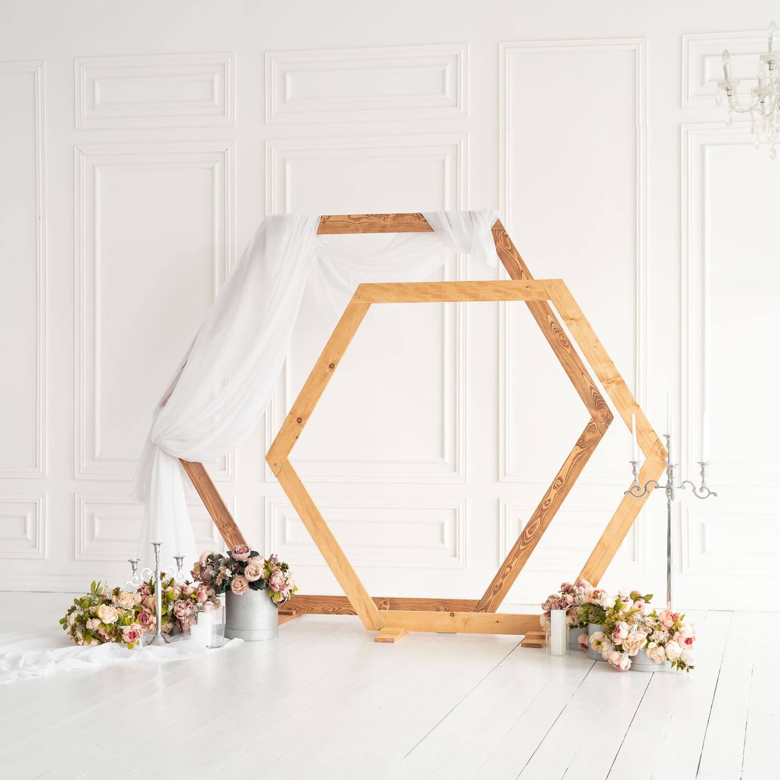 A hexagon-shaped wooden wedding arch decorated with white fabric and surrounded by floral arrangements.