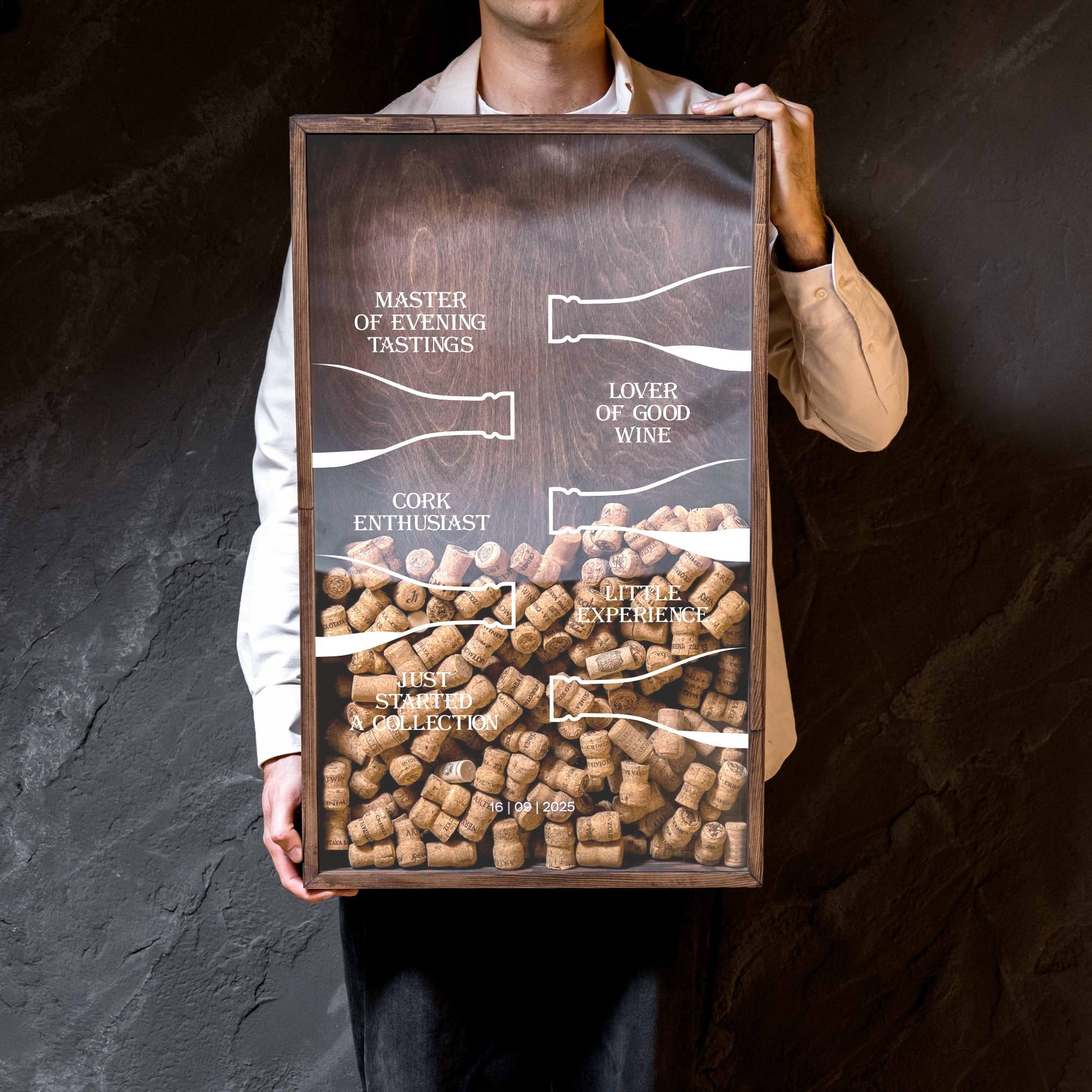 A framed art print featuring a graphic of wine corks with handwritten-style labels, held by a person wearing a white shirt and beige blazer against a dark textured wall.