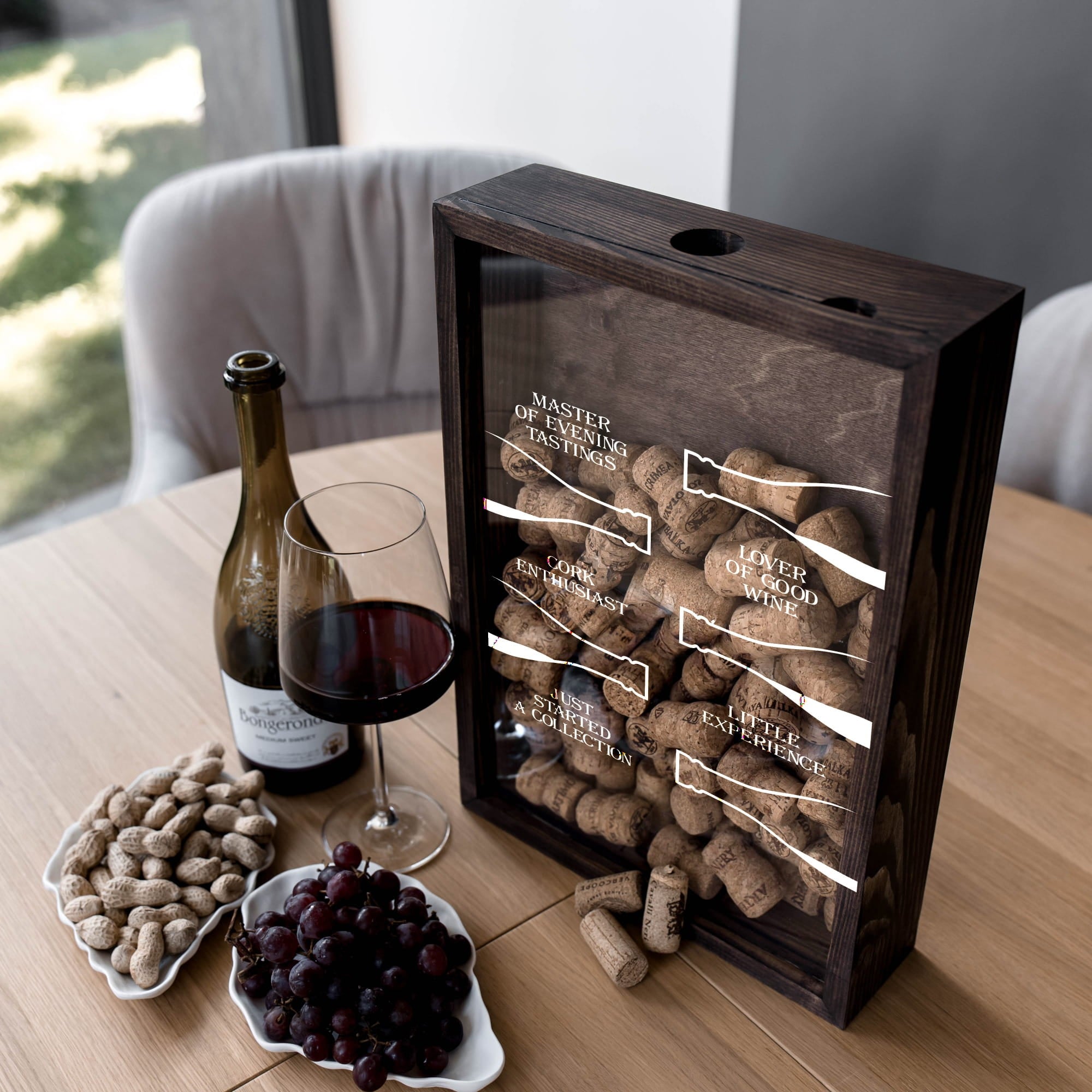 A dark wood wine cork display box with a glass front panel sits on a wooden table beside a bottle of red wine, a filled wine glass, peanuts, and grapes. The front shows labeled cork compartments.