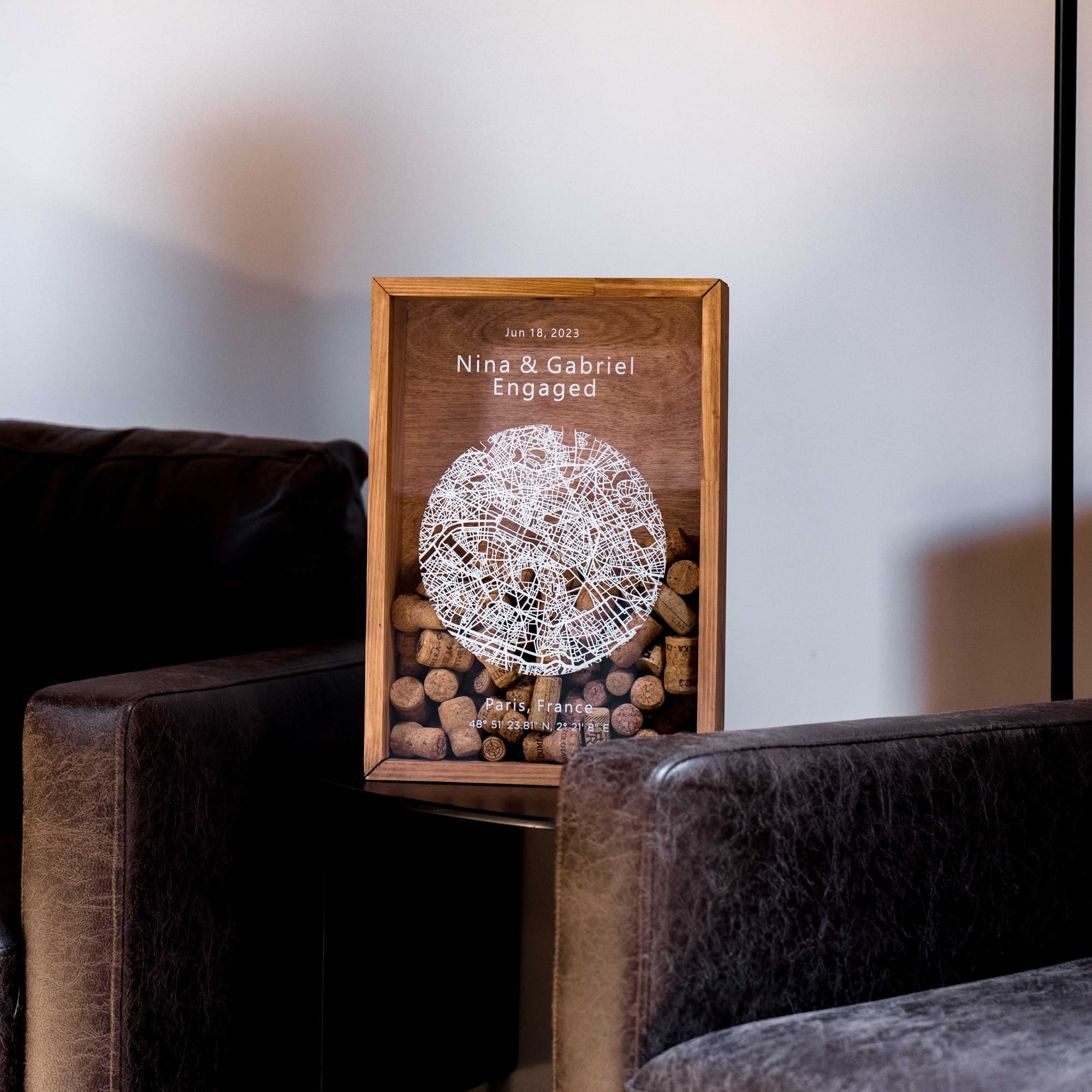 A framed wooden engagement gift box displayed on a side table between two dark leather armchairs.