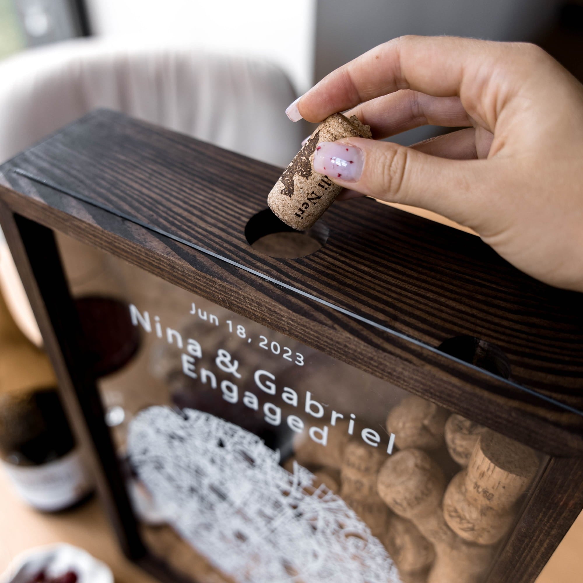 A close-up lifestyle shot of a hand placing a cork into a dark wood engagement box with a clear front panel.
