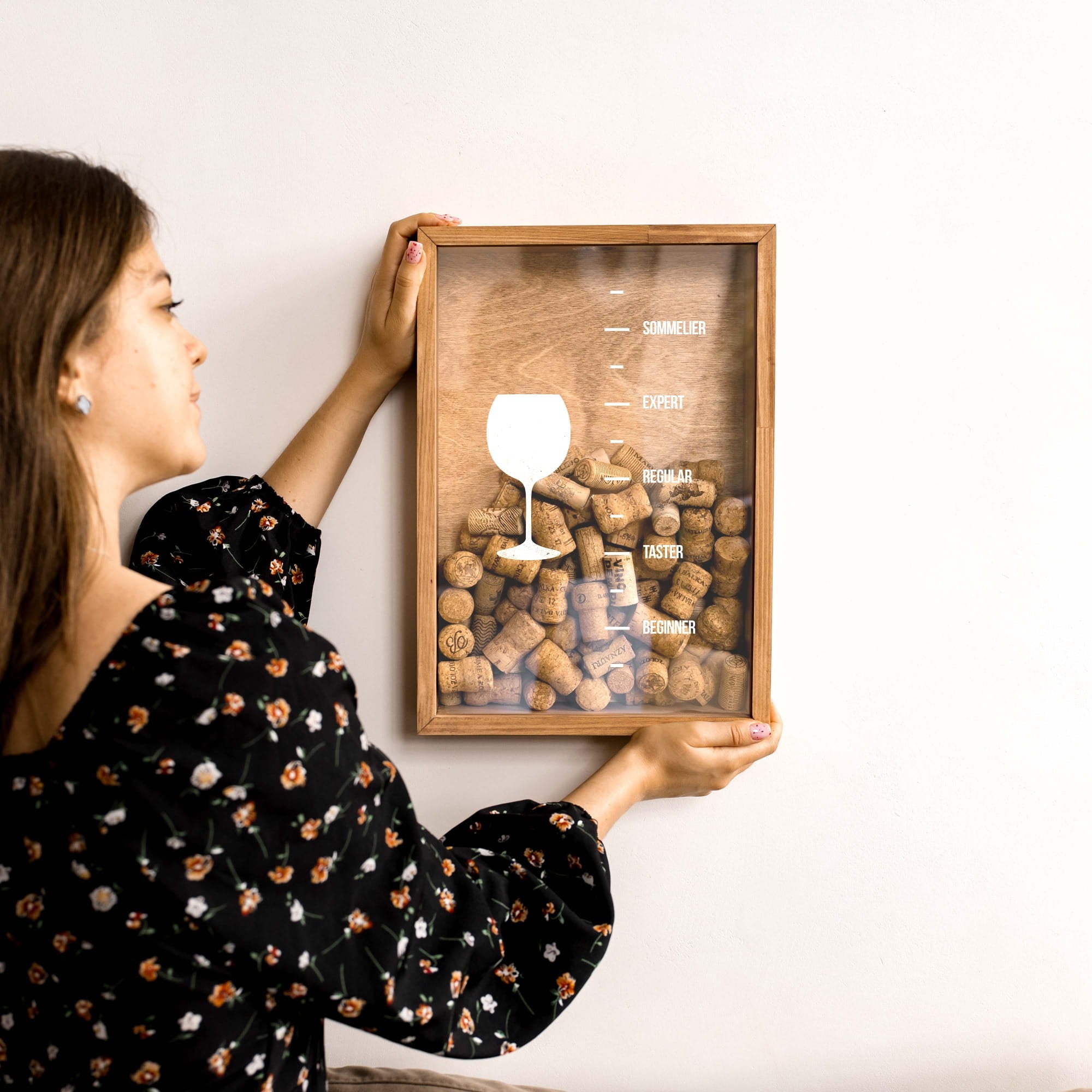 A woman in a black floral blouse holds a framed wall art piece against a plain white wall.