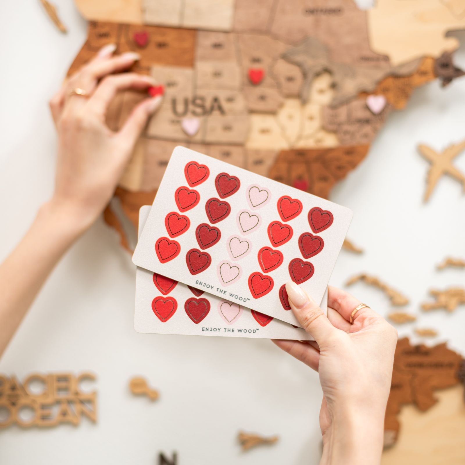 A person's hands holding a card with red heart-shaped pins, with a wooden map in the background.