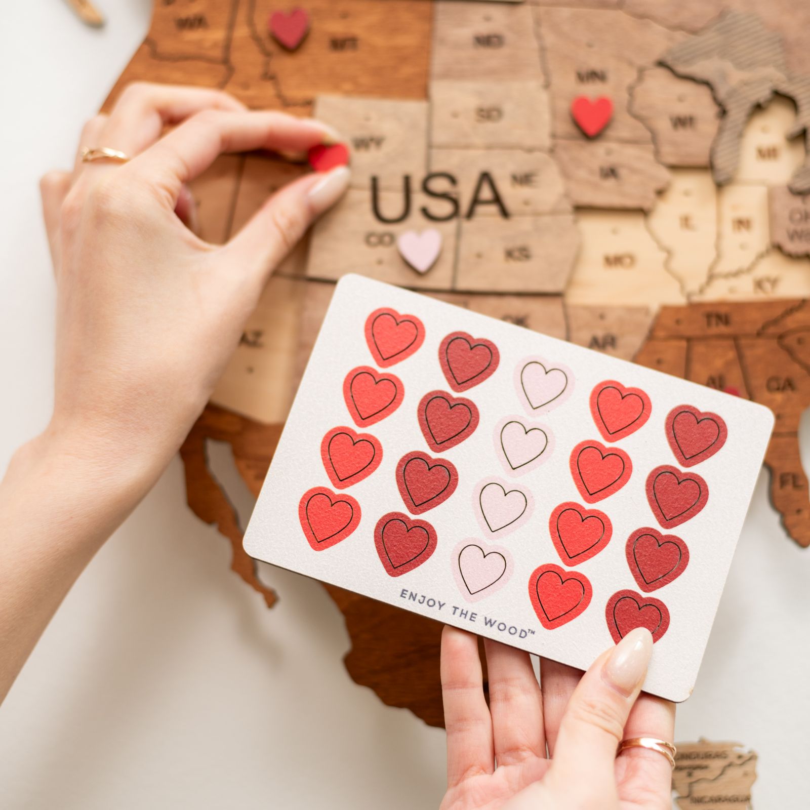 Hands placing red heart pin on wooden USA map with Enjoy The Wood travel accessories