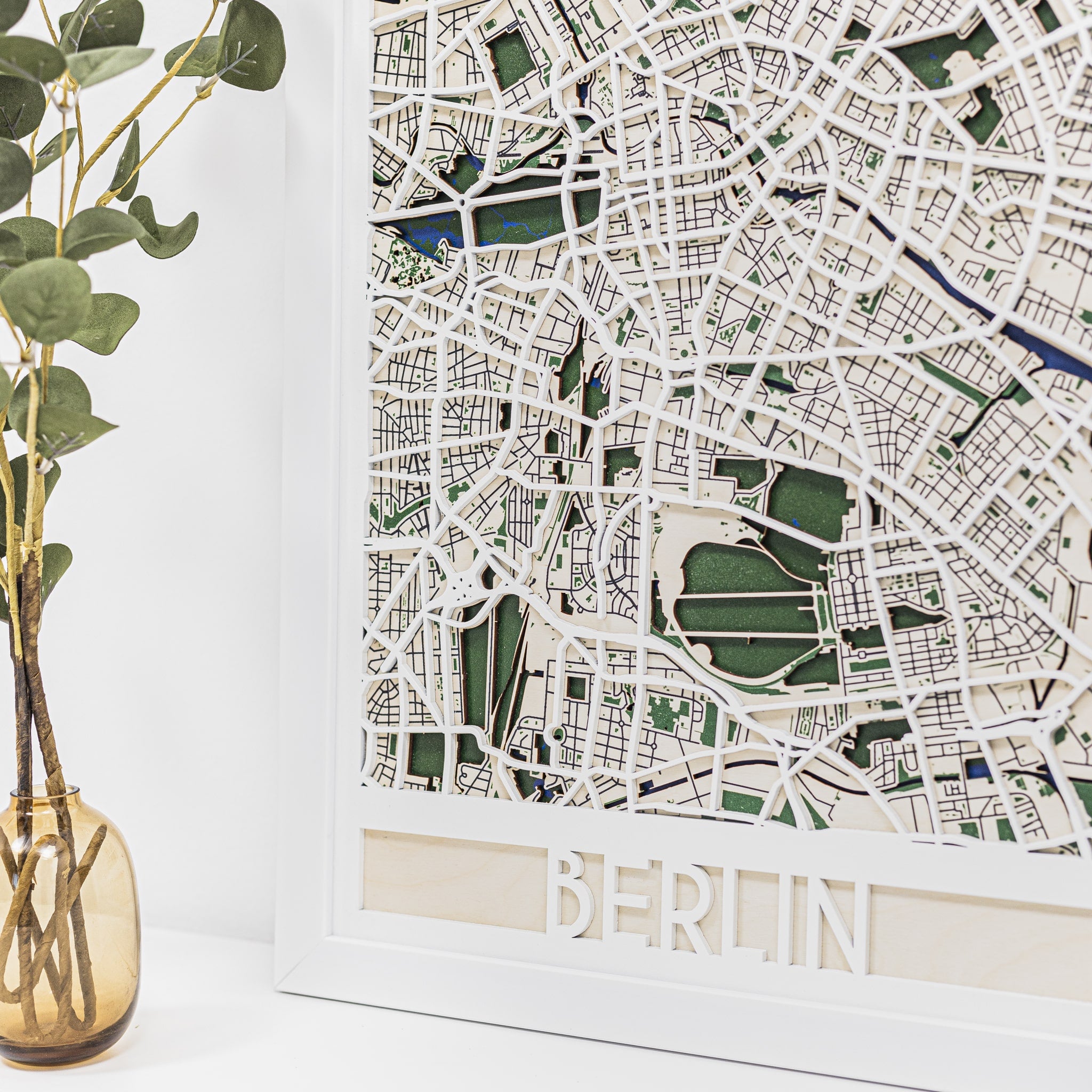 Wooden cutout map of Berlin with 'BERLIN' engraving, next to a vase with greenery on a white background