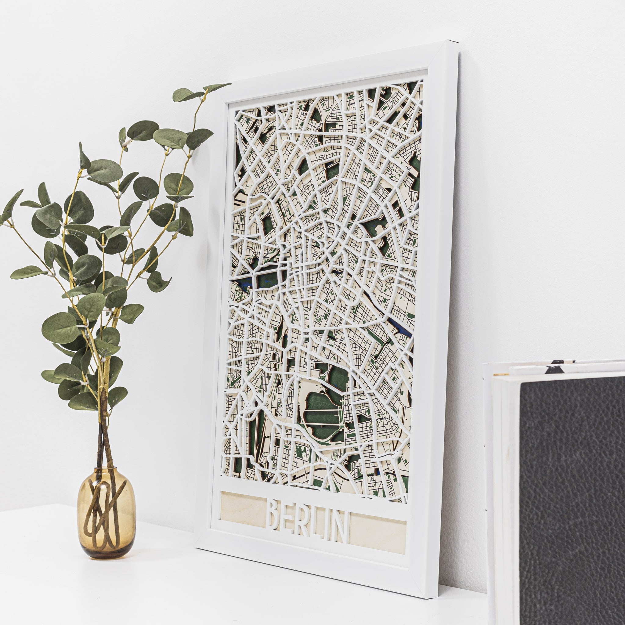 Framed city map of Berlin on a white wall with a plant and black box in the foreground.