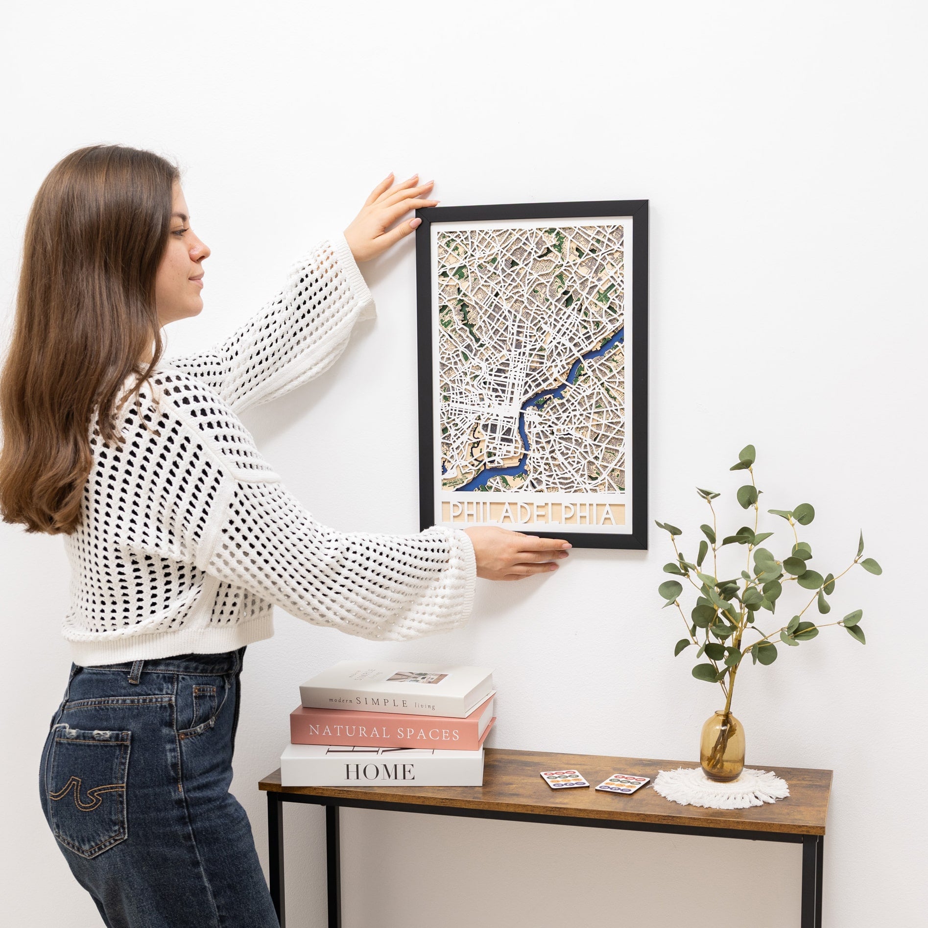 Woman hanging a framed map on a wall next to a small table with books and a plant.