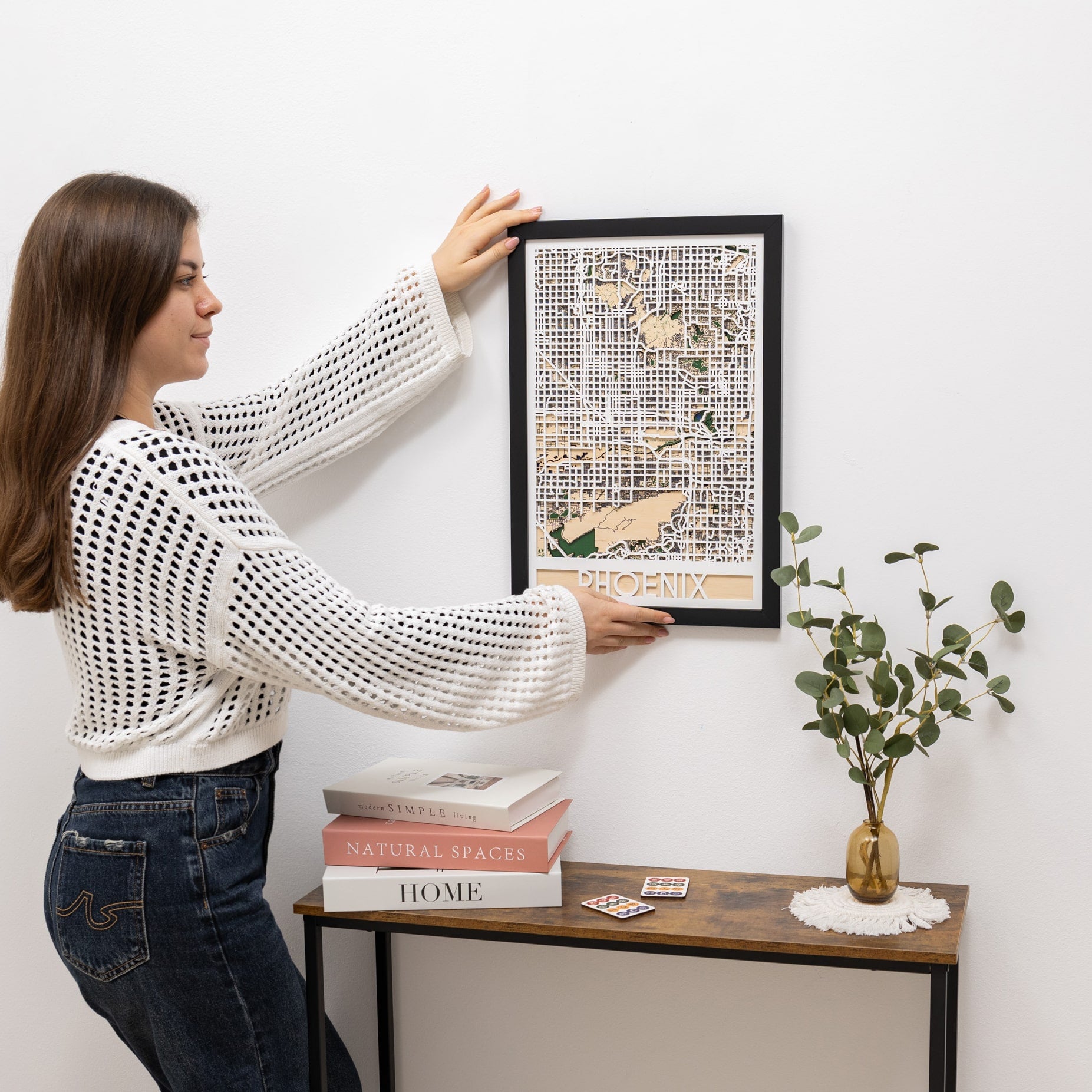 Woman hanging a framed artwork on a wall next to a small table with books and a plant.