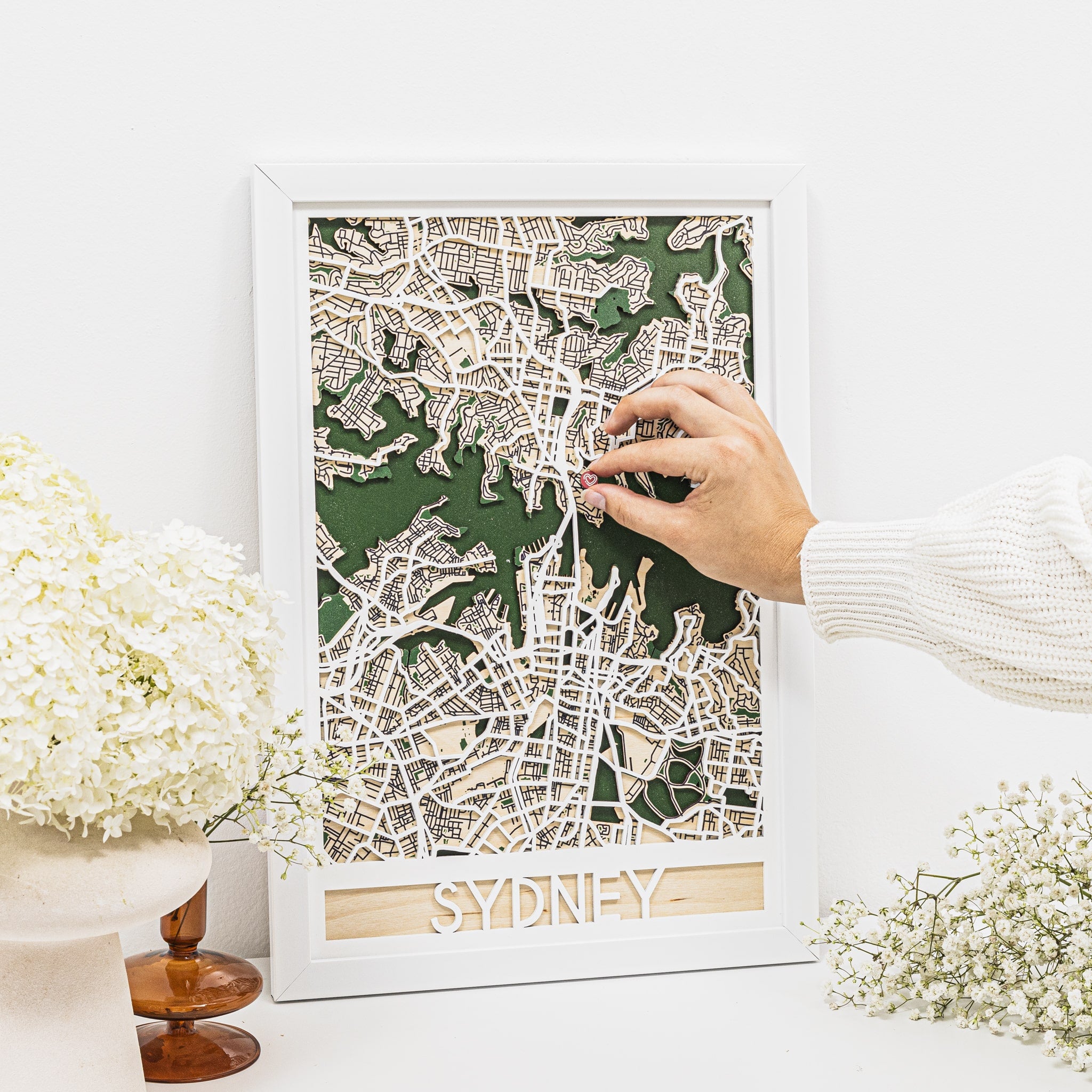 Framed map of Sydney with a hand adjusting it on a white surface with decorative elements.
