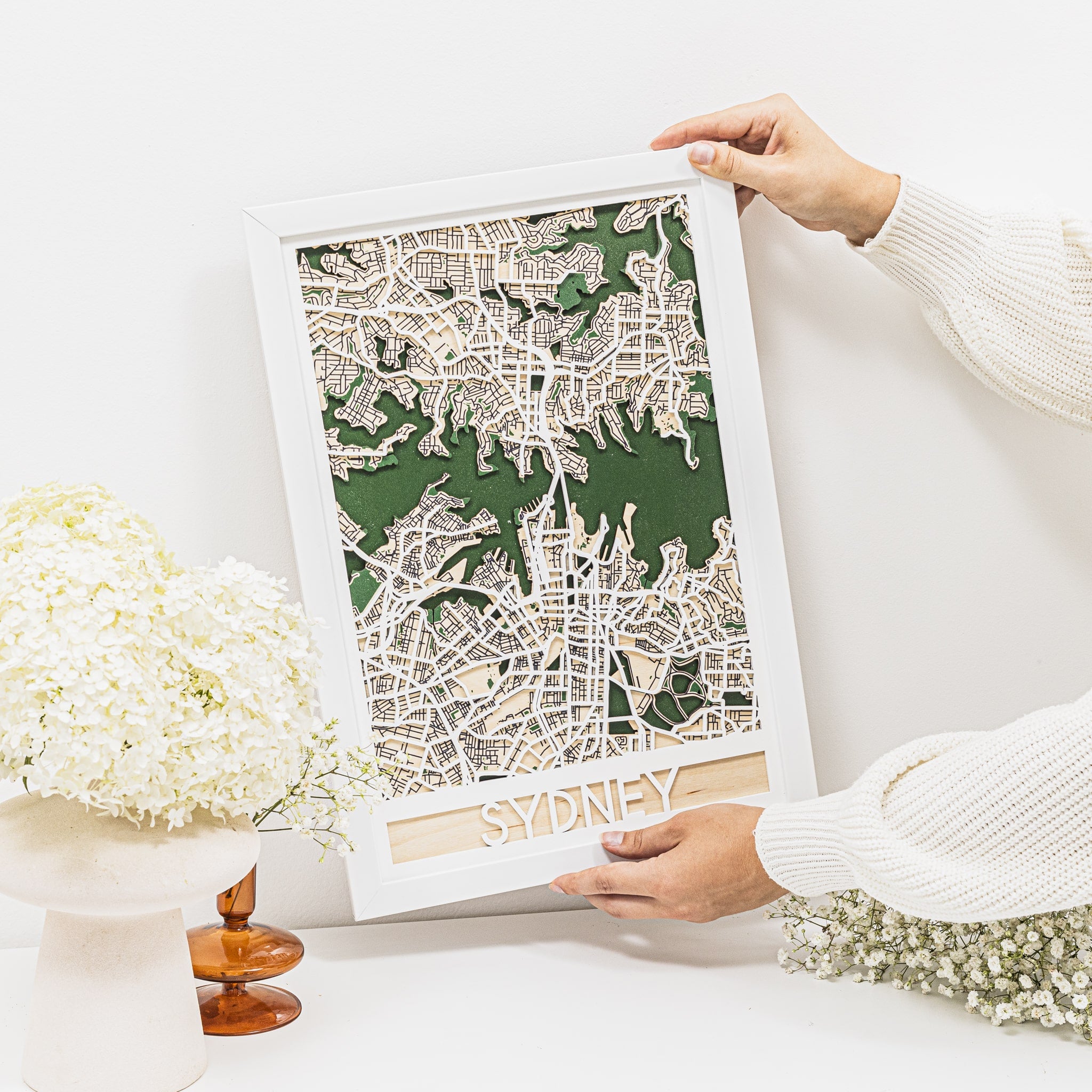 Person holding a framed map of Sydney on a white surface with flowers in the background