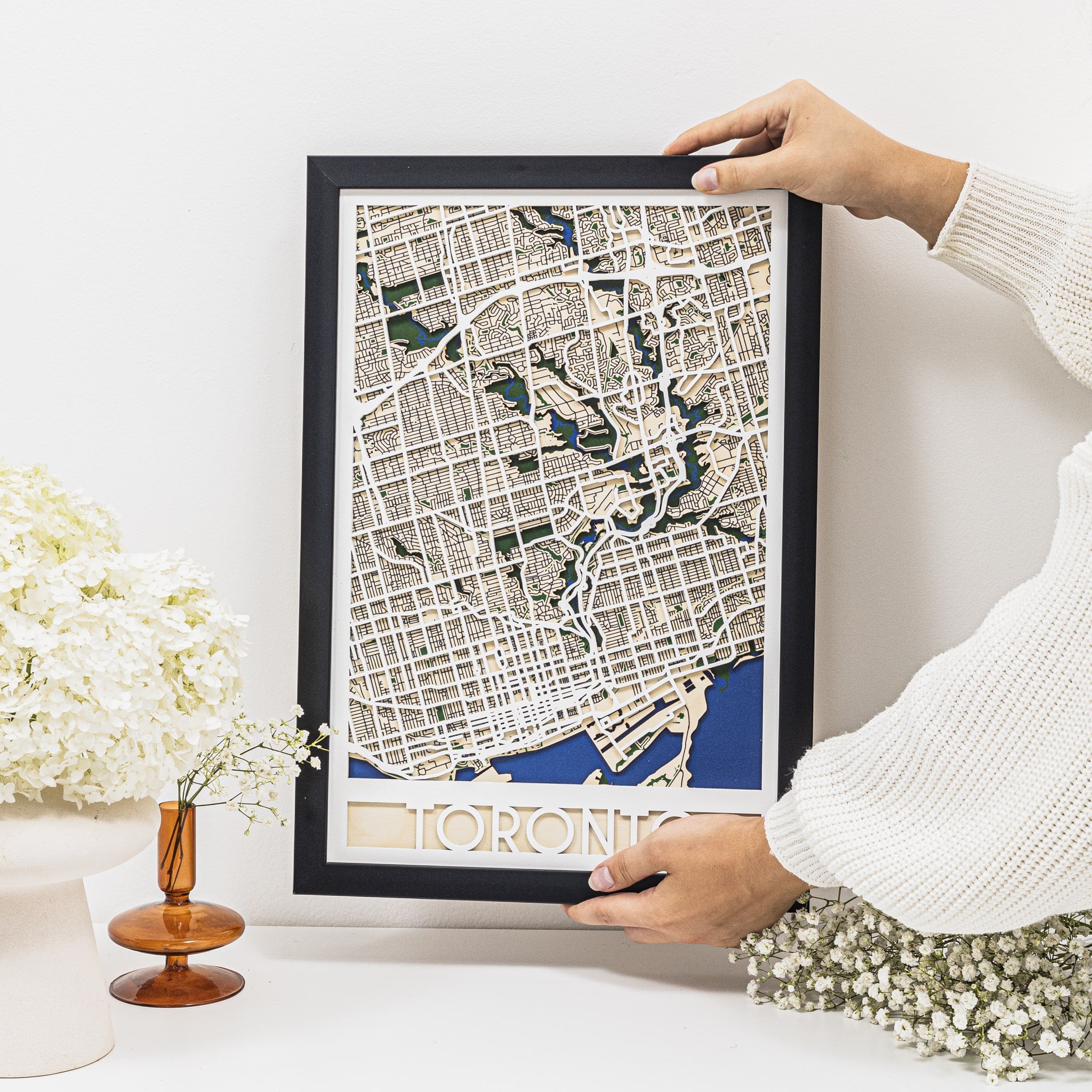 Framed map of Toronto held by a person in a home setting with decor elements.