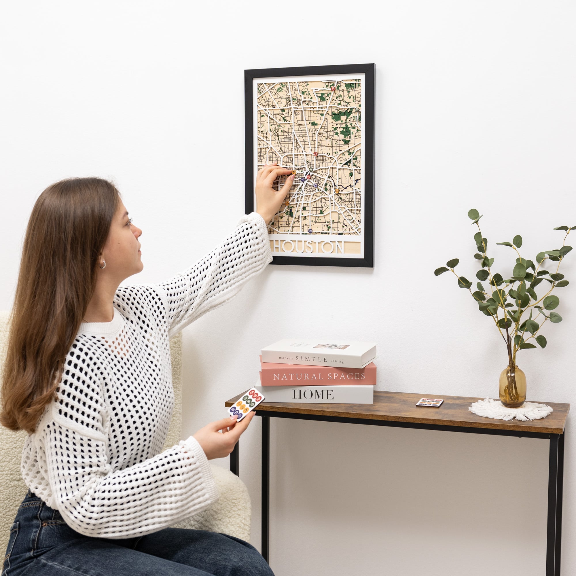 Woman hanging a framed map on a wall next to a wooden side table with books and a plant.