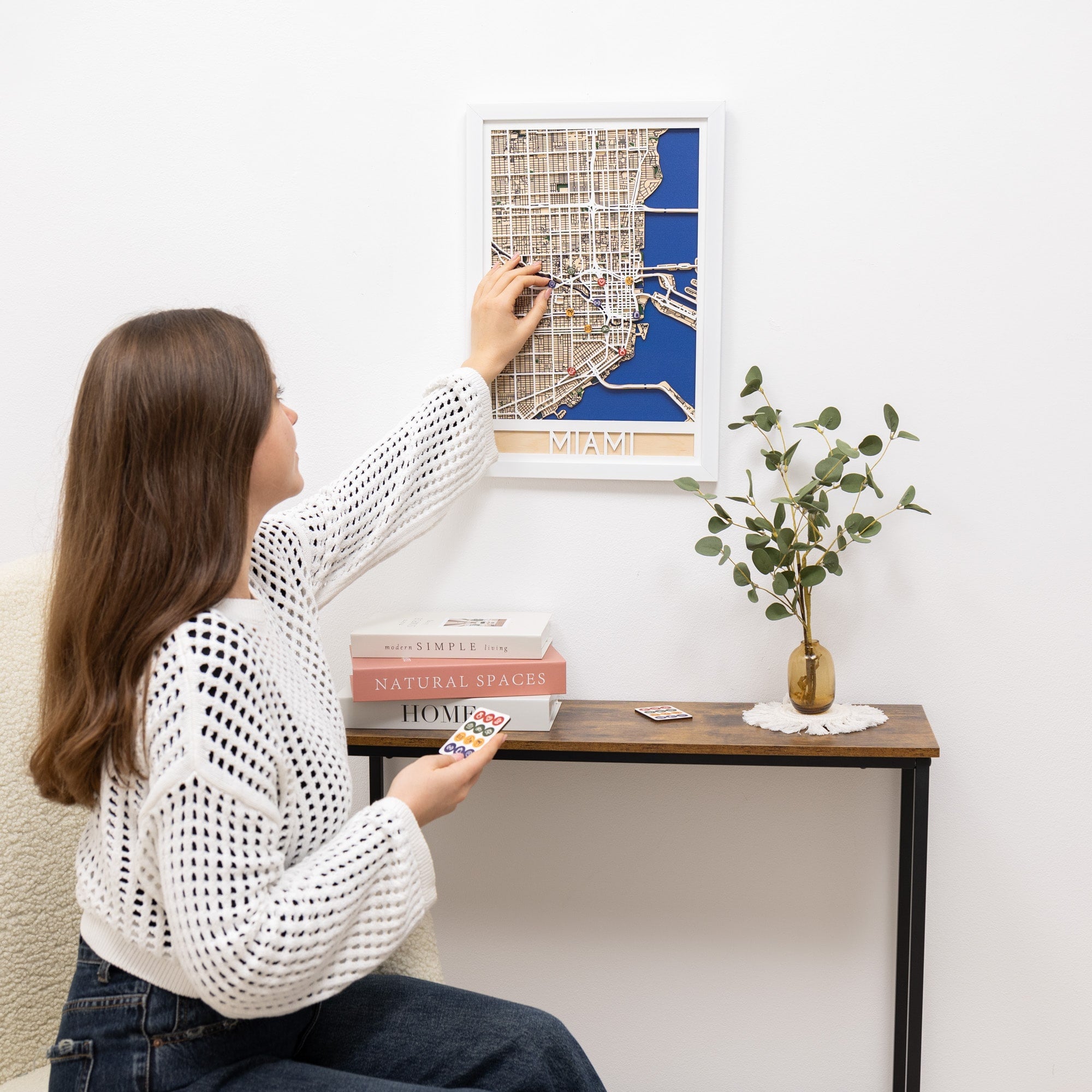 Woman hanging a framed map on a wall next to a table with decor items.