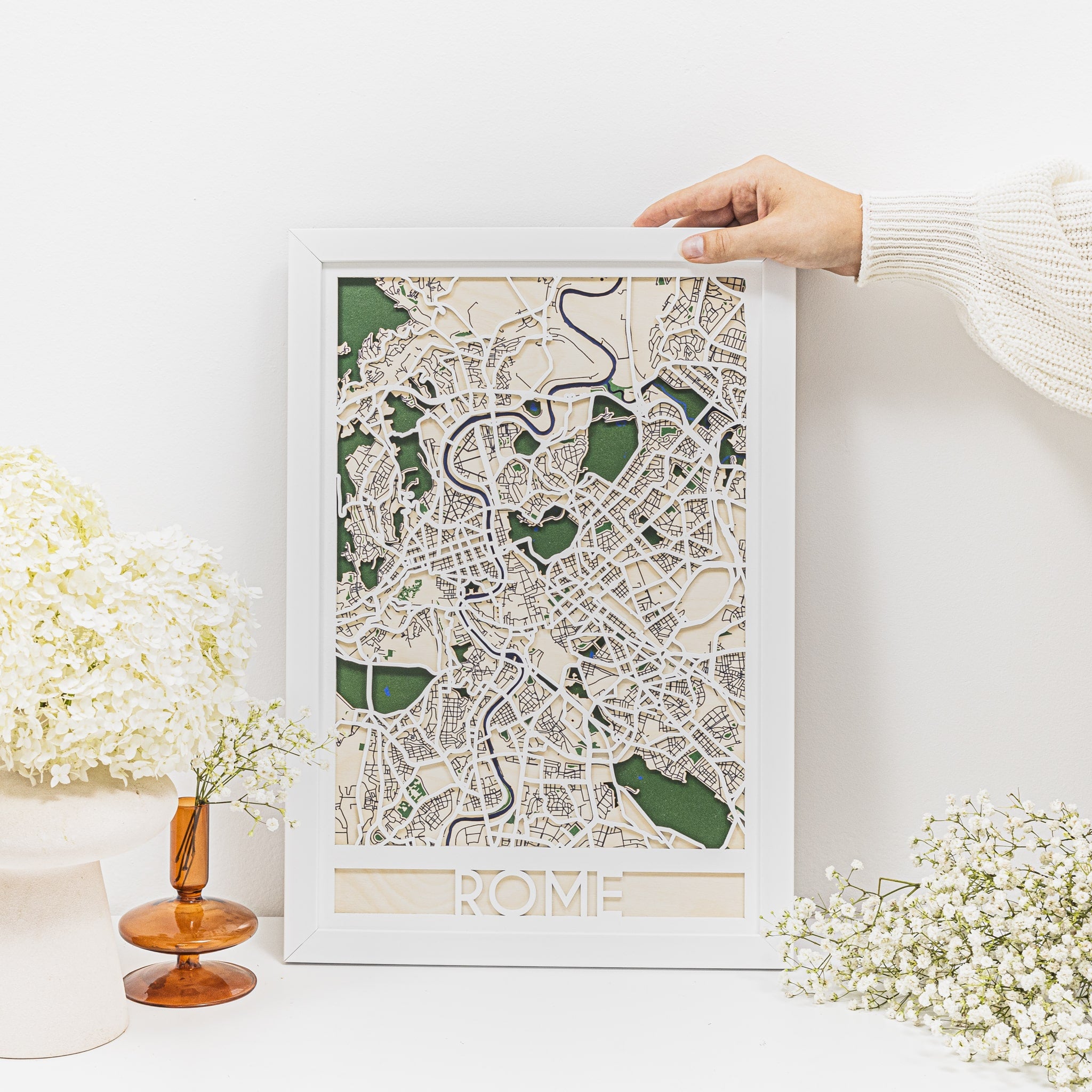 Framed map of Rome held by a person on a white surface with decorative elements.