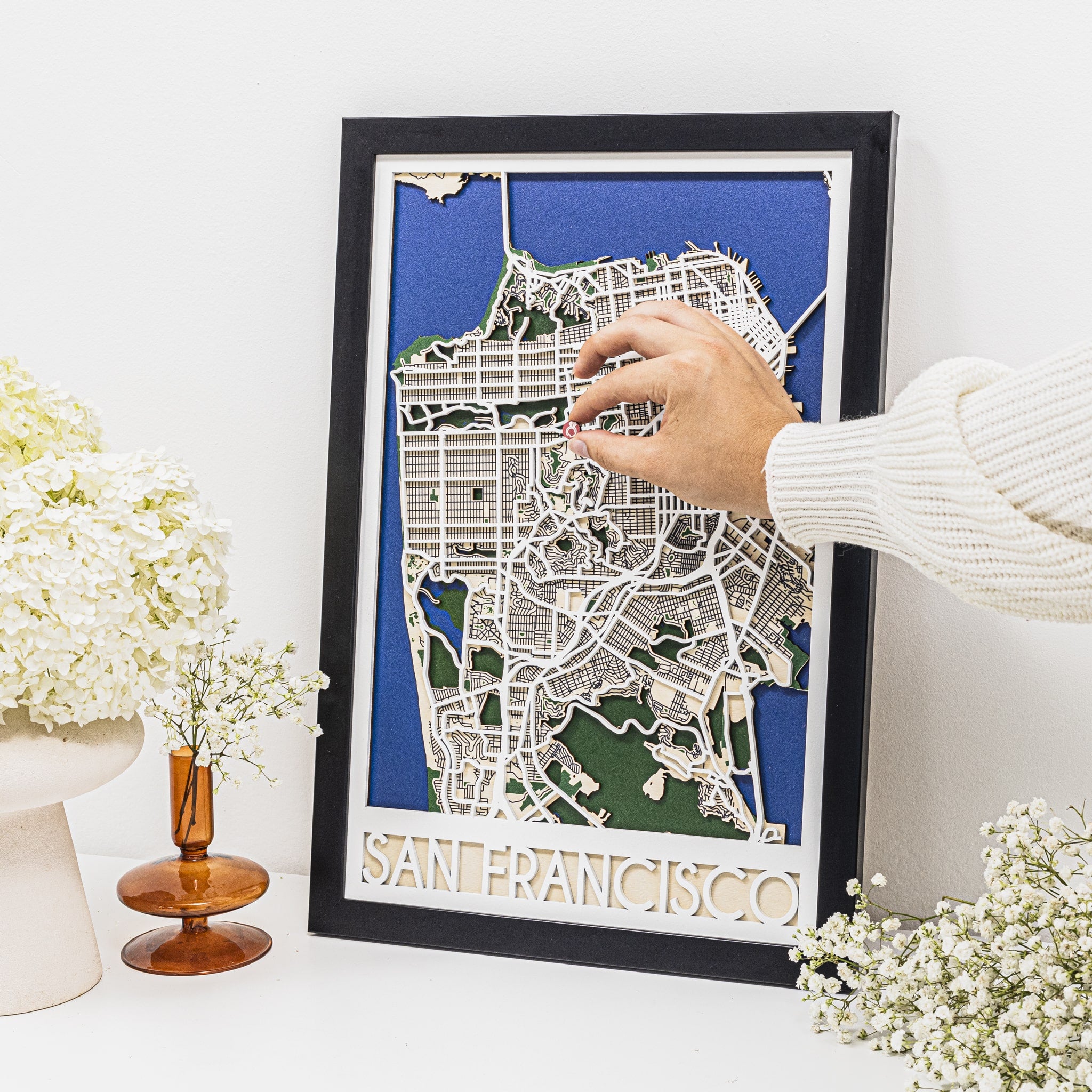 Framed map of San Francisco with a hand touching it, surrounded by decorative flowers on a white surface.