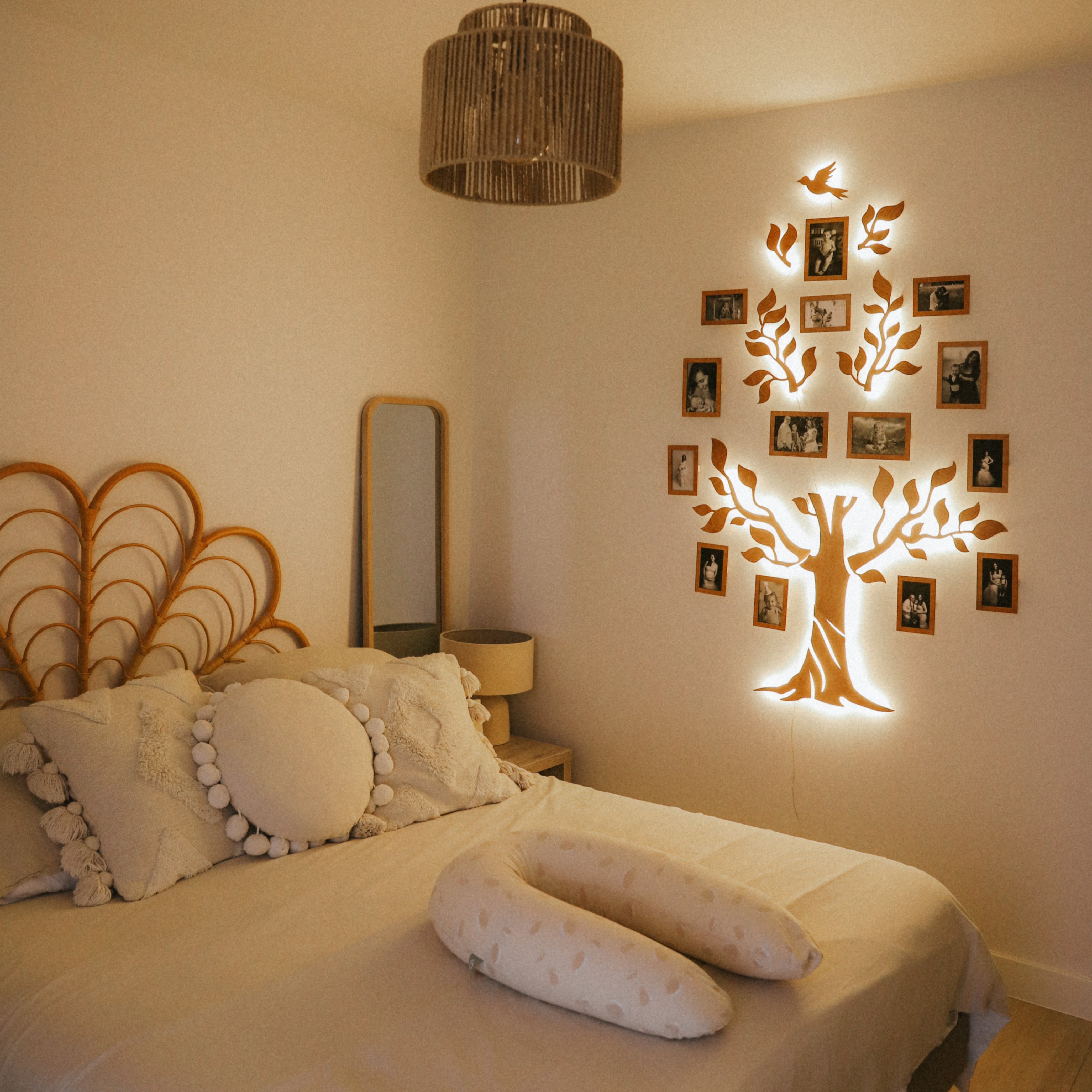 A wooden LED family tree decor mounted on a wall above a bed, with LED lights illuminating its branches and leaves, and photo frames attached to its branches.