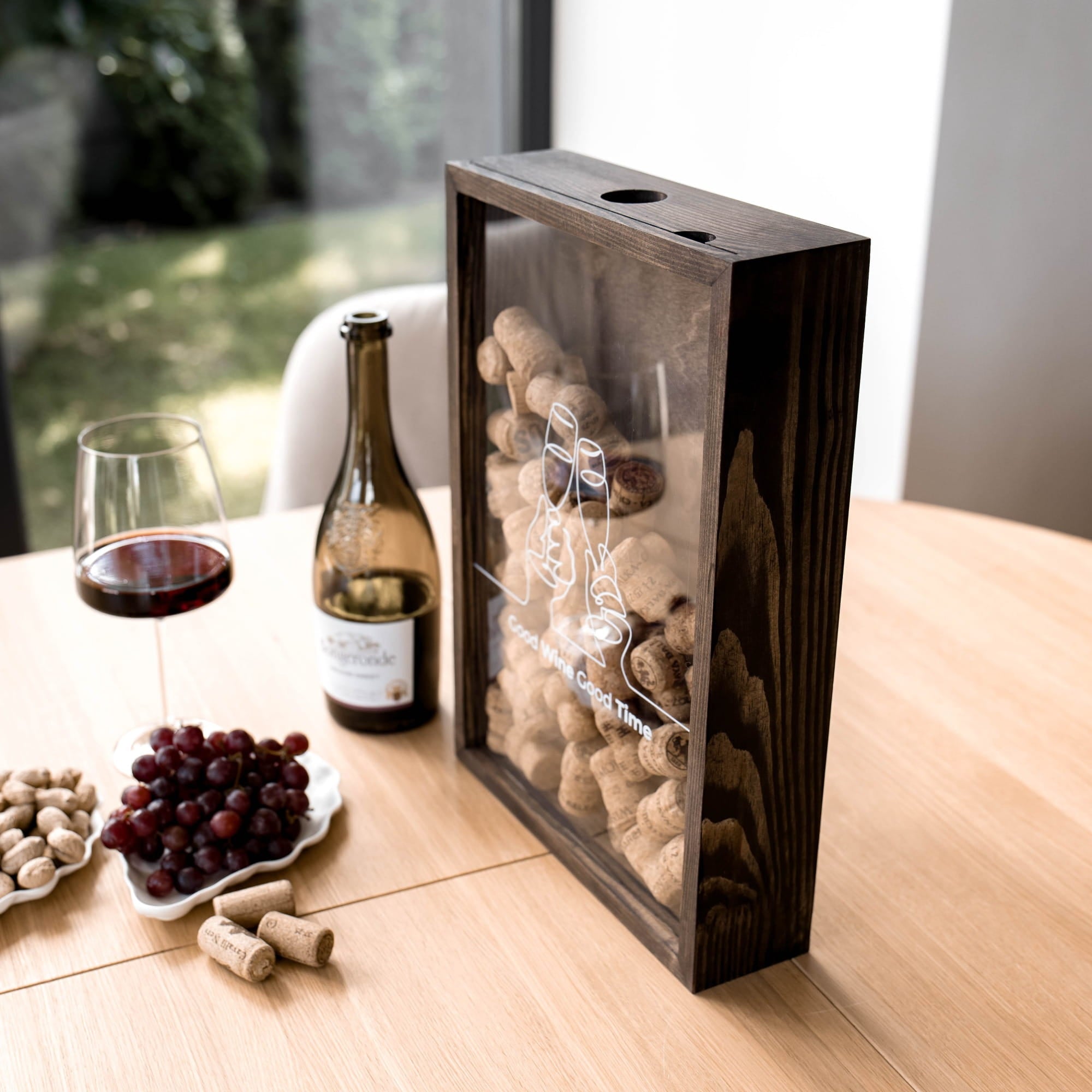 A dark wooden wine cork display box with a clear front panel stands on a light wood table beside a bottle of wine