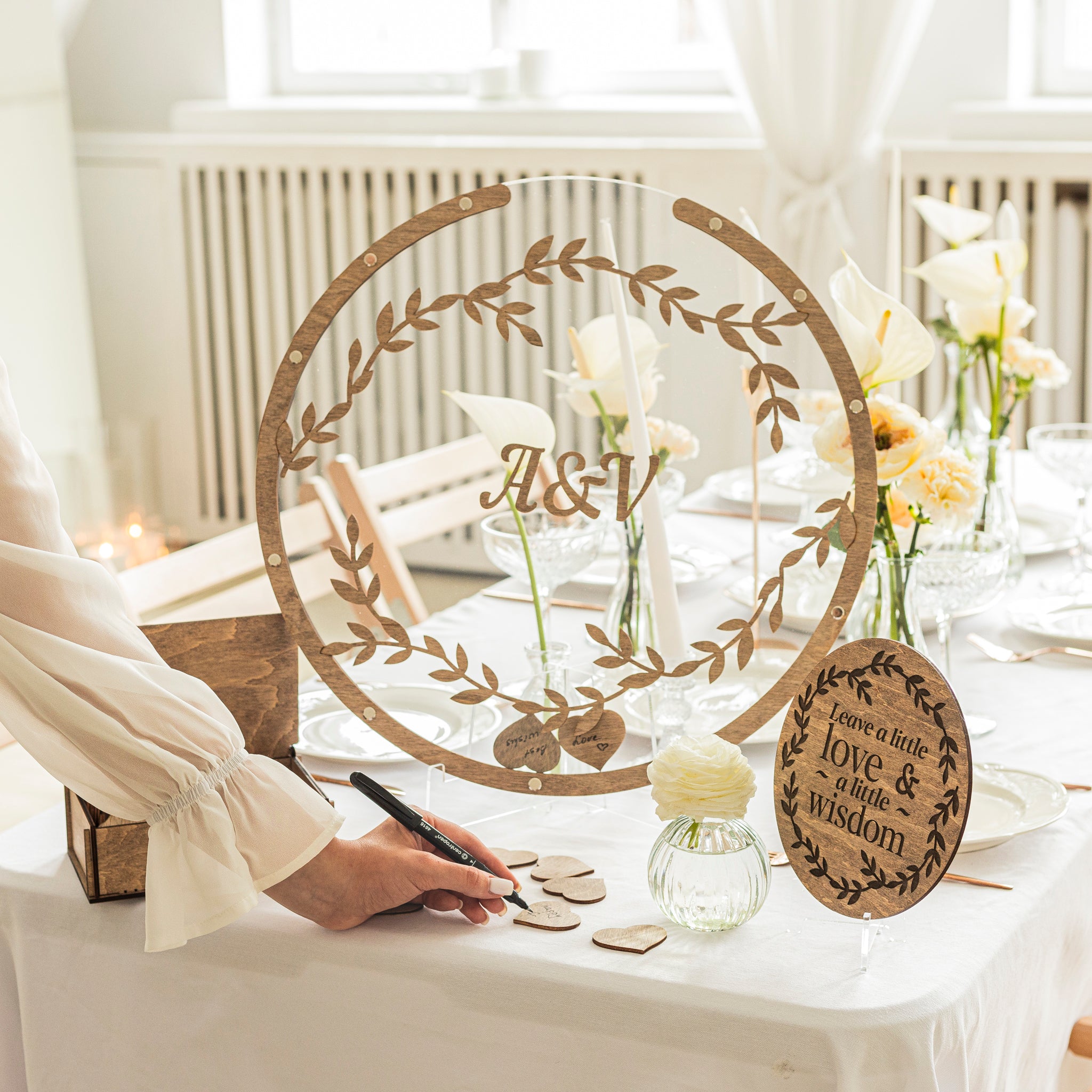 Decorative table setting with a circular wooden piece featuring initials and floral design, surrounded by flowers and a person writing.