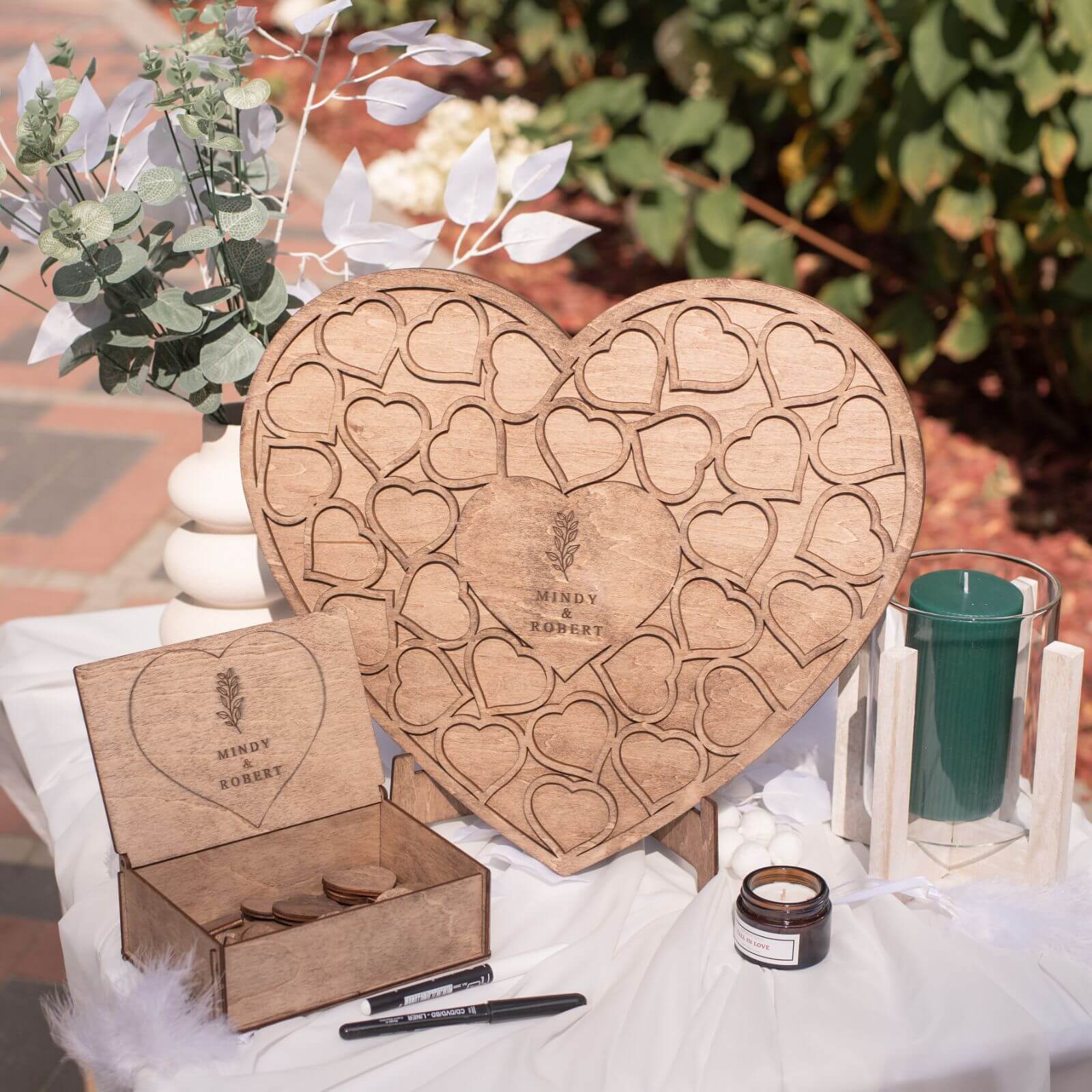 A heart-shaped wooden guest book with cut-out spaces for writing, placed on a table with decorative elements around it.