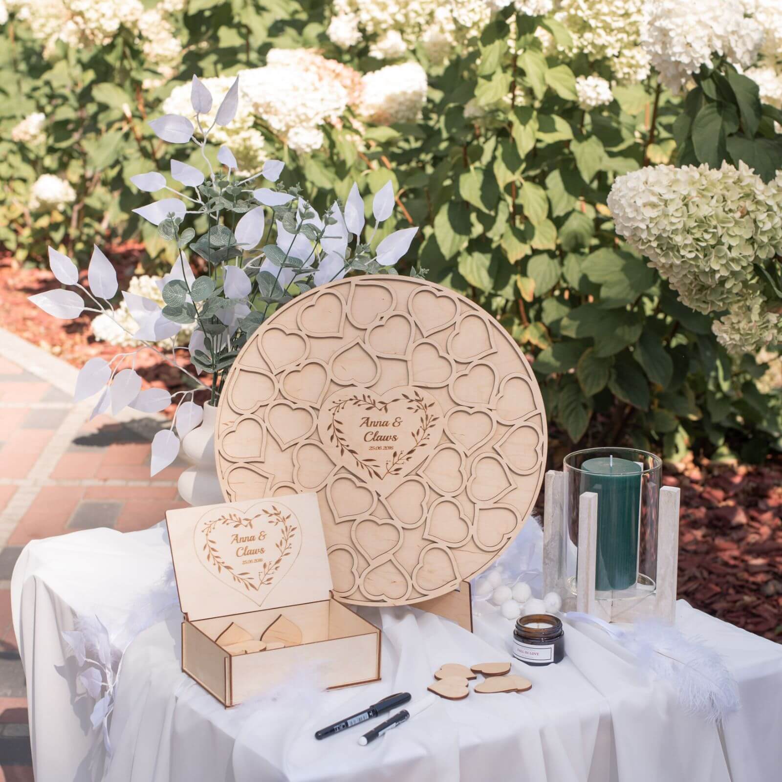 A wooden heart-shaped wedding guest book alternative set on a table, with a pen and marker provided for writing messages. The background features greenery, indicating an outdoor setting.