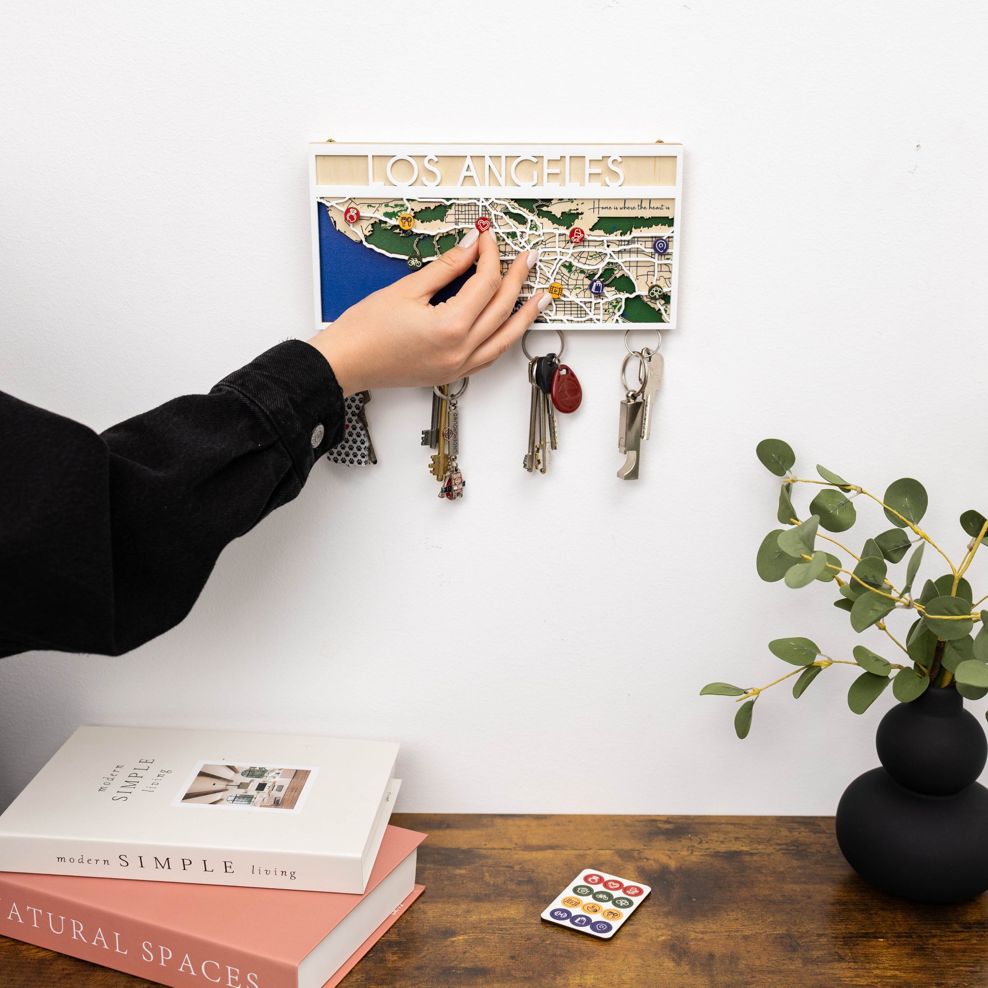 Person placing keys on a magnetic key holder with a map of Los Angeles, books, and a plant in the background.