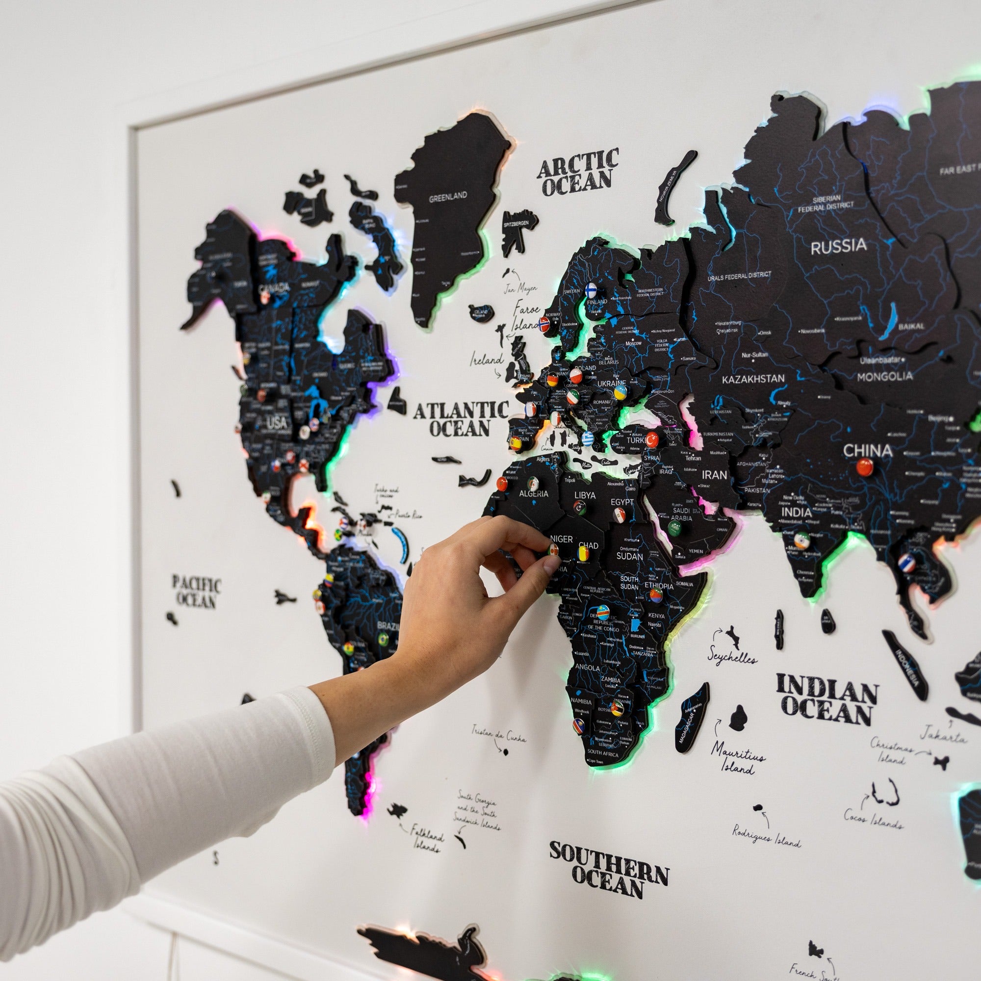 World map with colorful pins on a white wall, showing a hand interacting with it.