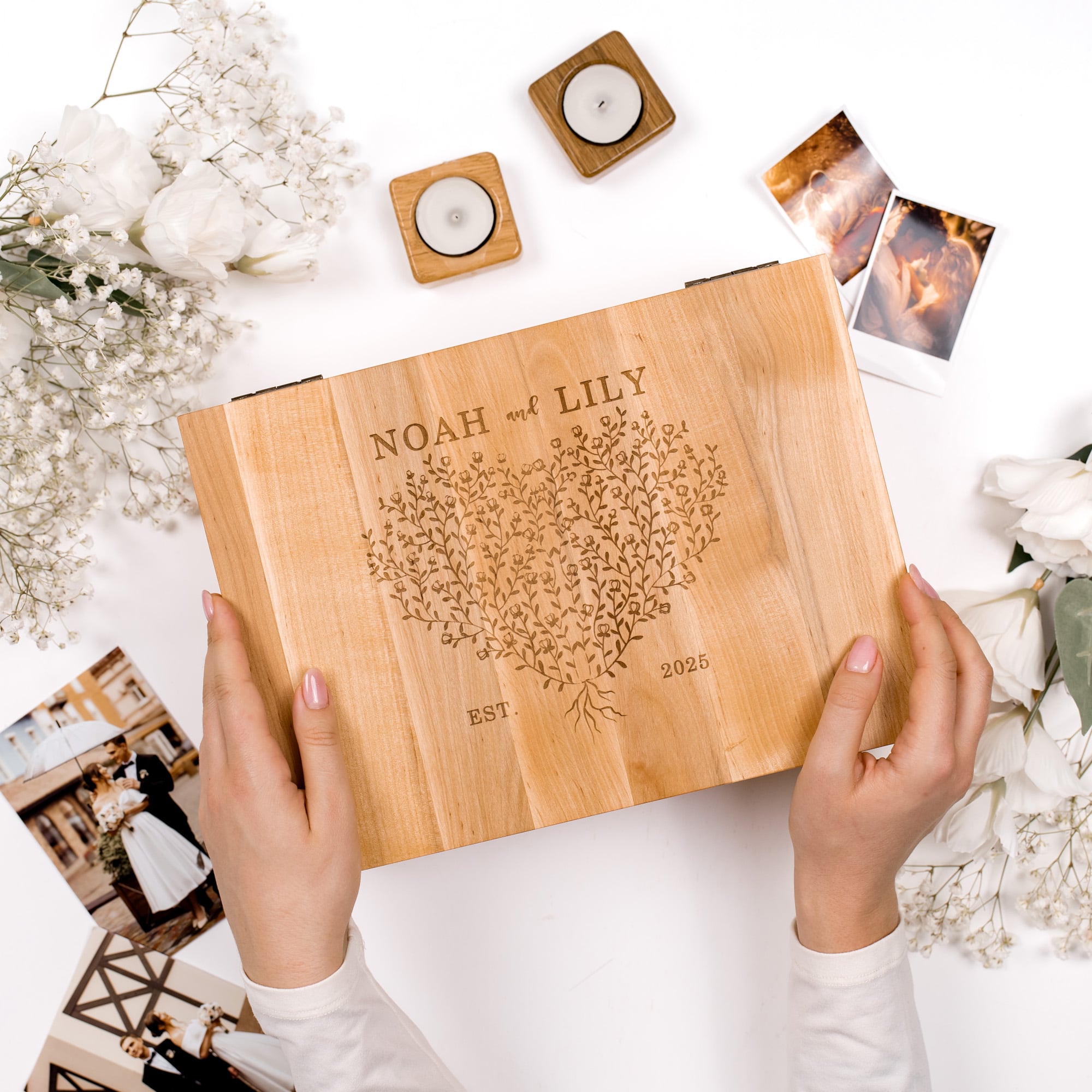 Wooden album with engraved text held by hands on a white surface with flowers and photos.