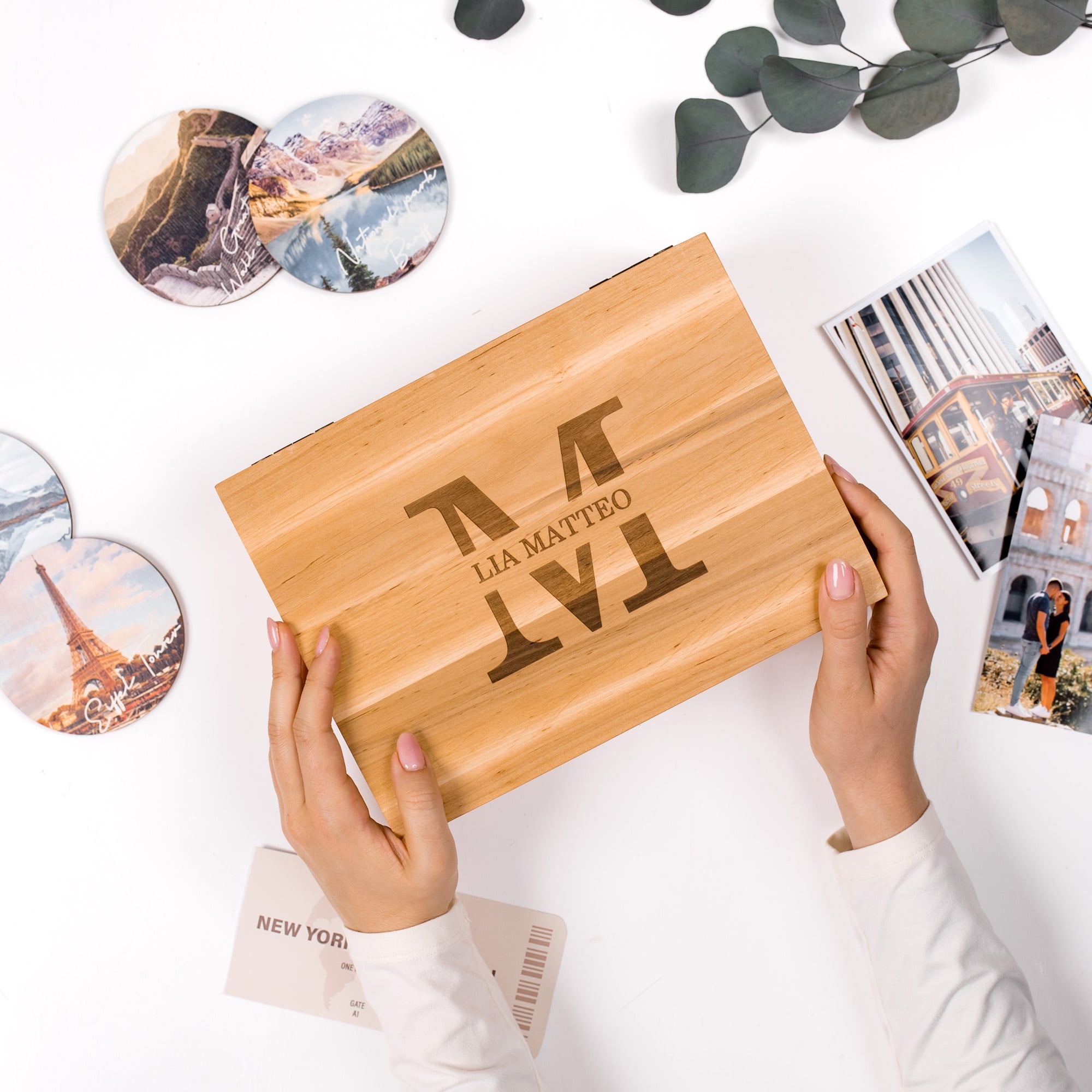 Wooden box with engraved text held by hands on a white surface with travel-themed items.