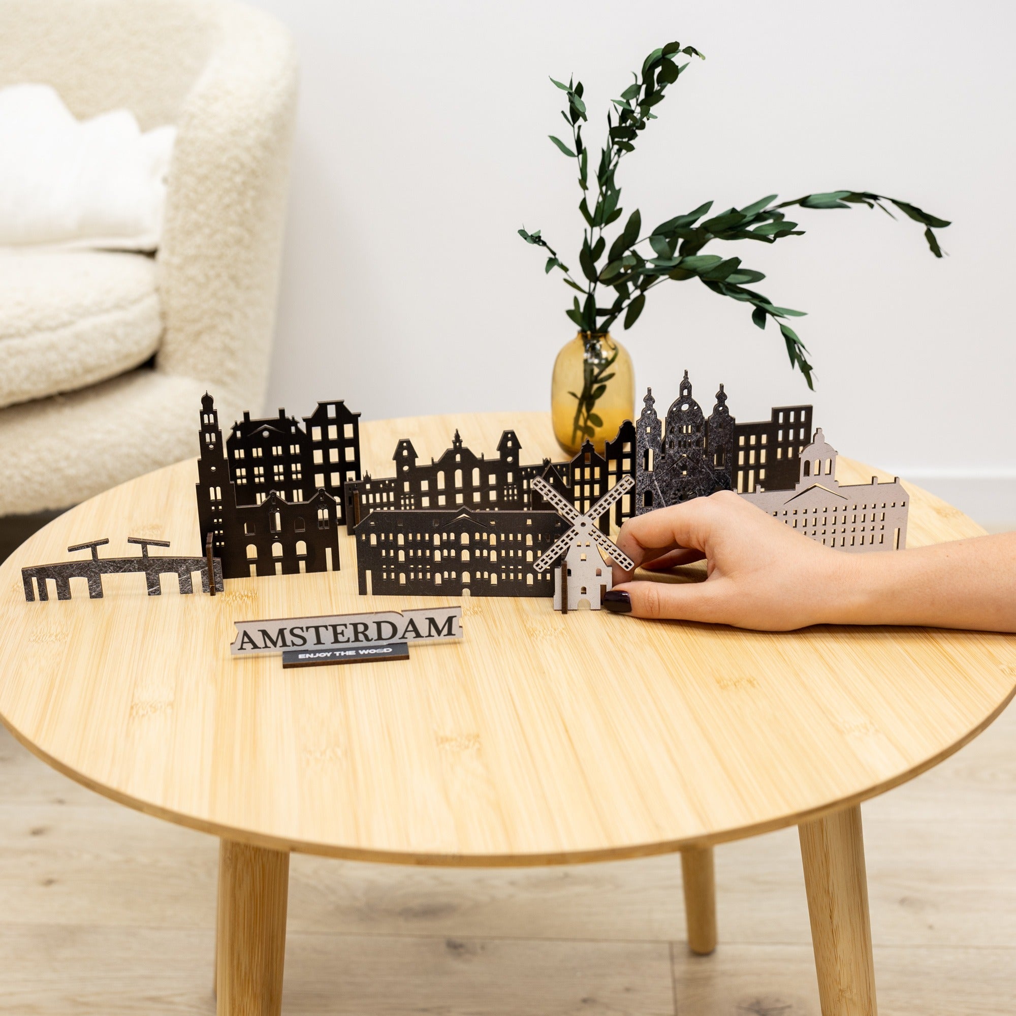 Model of Amsterdam skyline on a wooden table with a hand holding it, next to a plant and 'Amsterdam' sign.