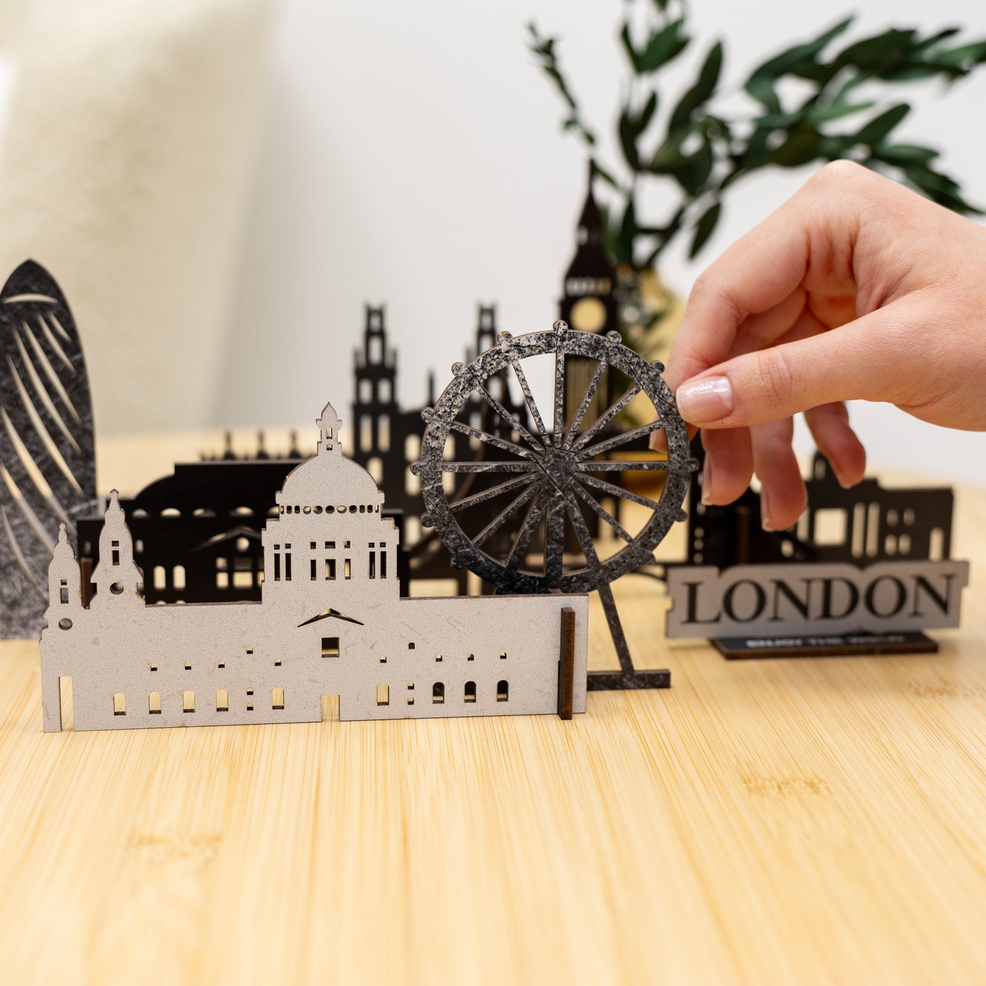 Hand holding a metal model of the London Eye with other London-themed miniatures on a wooden surface.