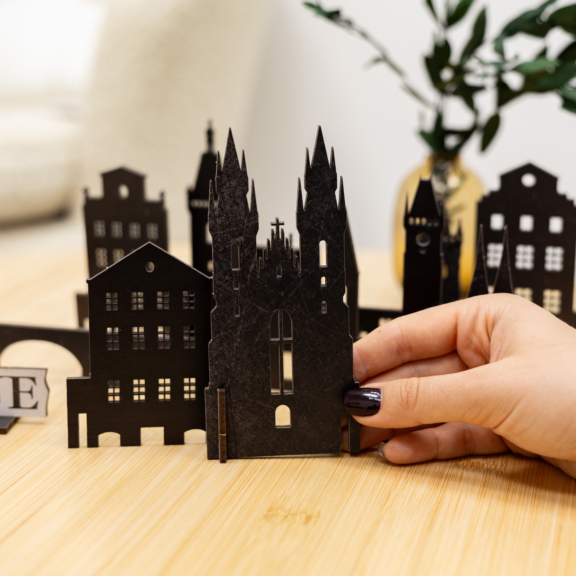 Hand holding a black paper model of a castle on a wooden surface with other similar models in the background.