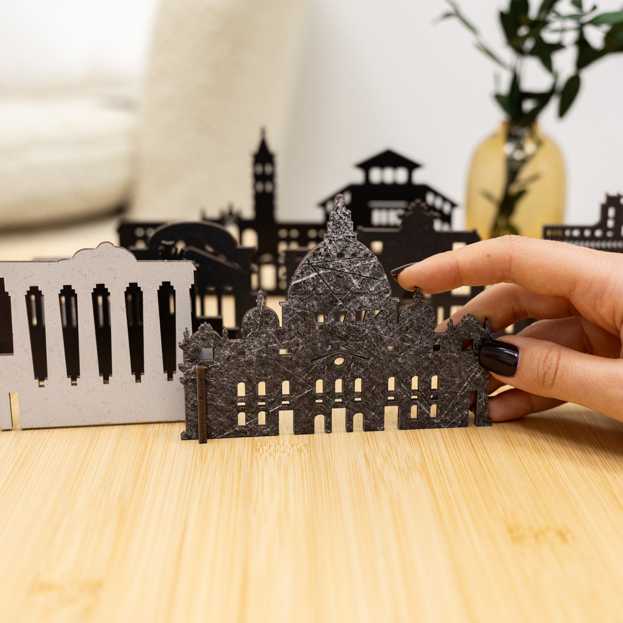 Hand holding a cut-out architectural model of a building on a wooden surface with a blurred cityscape in the background.