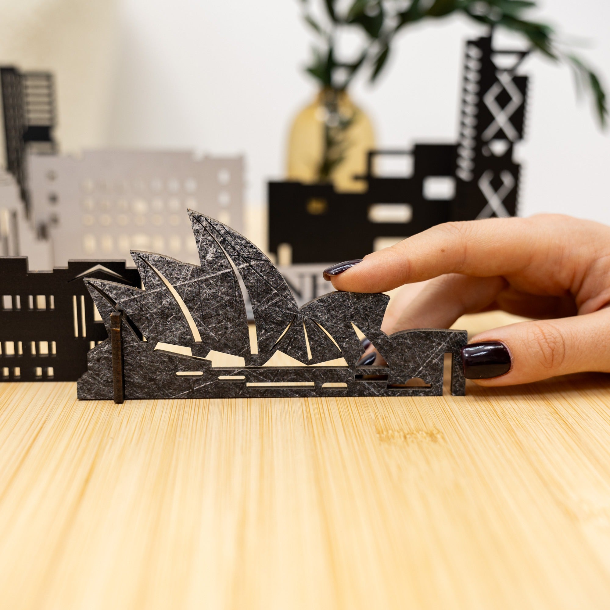 Hand holding a miniature model of the Sydney Opera House against a blurred cityscape background.