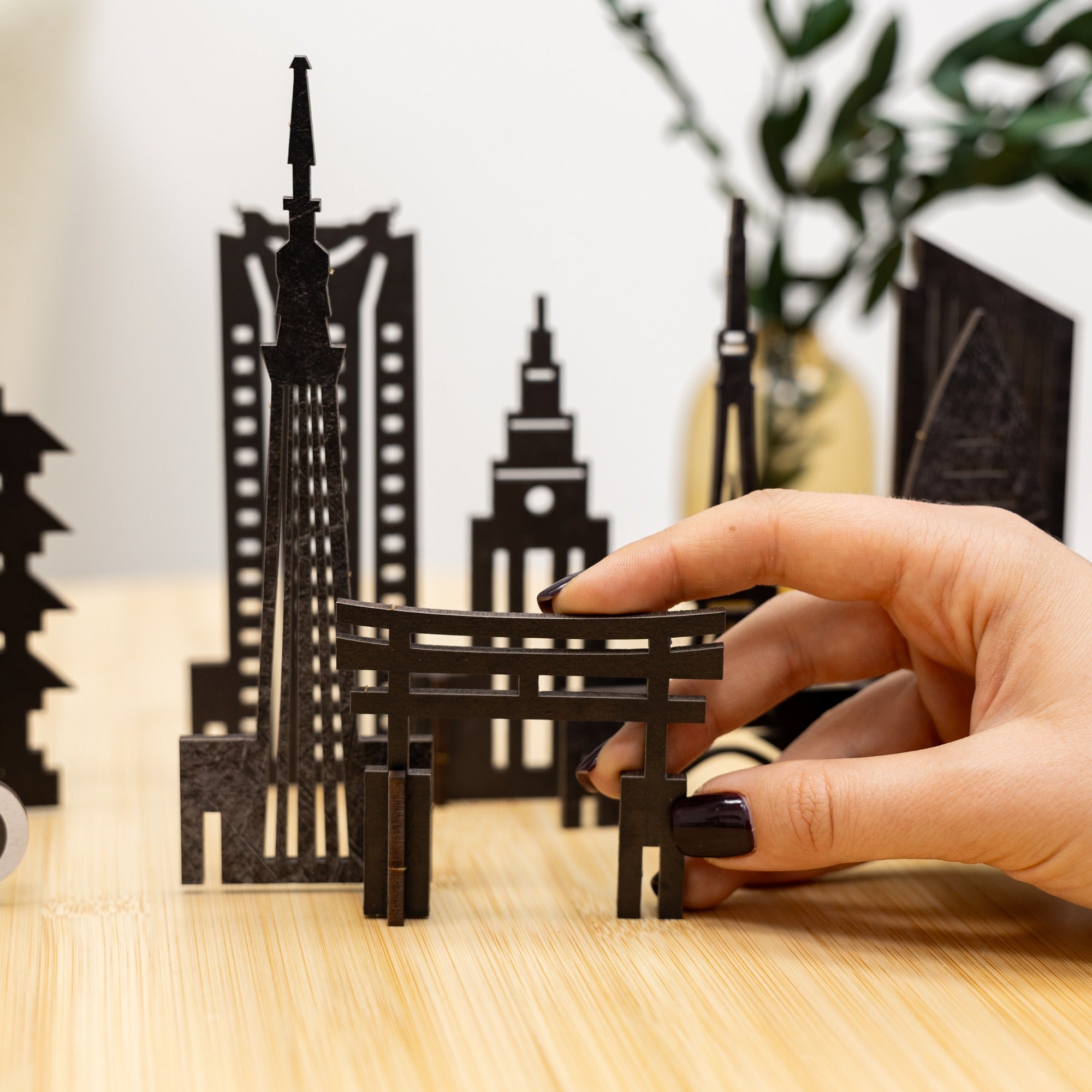 Hand arranging black paper models of skyscrapers on a wooden surface with a blurred plant in the background