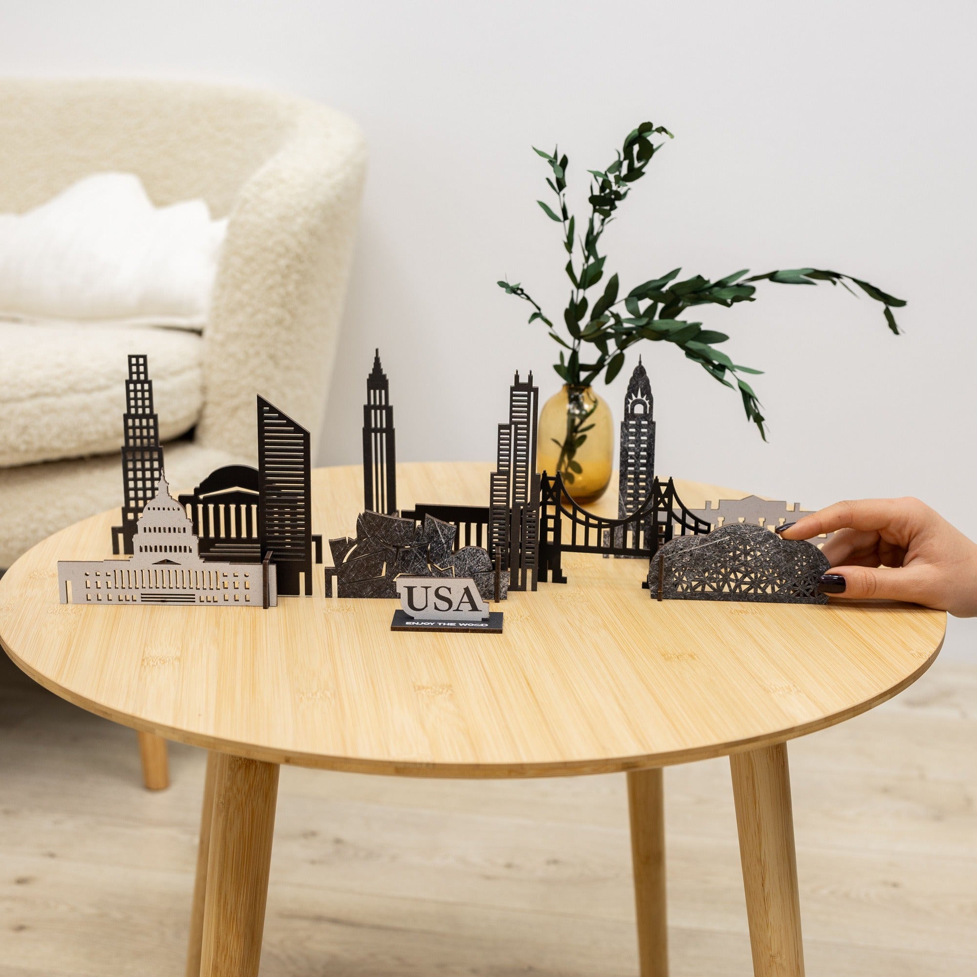 Hand holding a miniature cityscape model on a wooden table with a plant and sofa in the background.