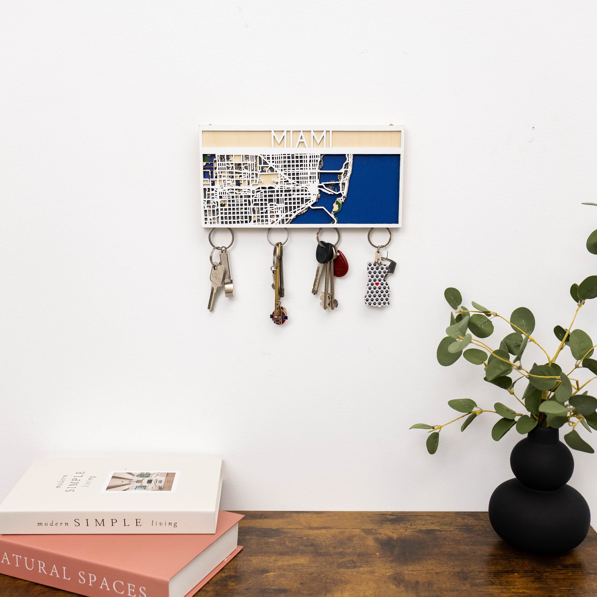 Key holder with keys on a wall above a desk with books and a vase.