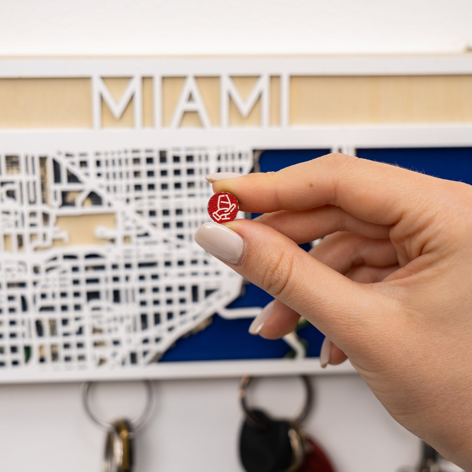 Hand holding a red and white keychain in front of a decorative wall piece with 'Miami' on it.