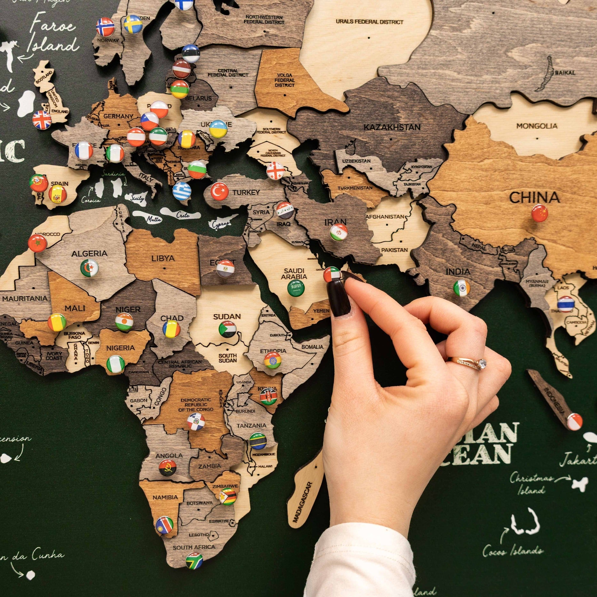 Wooden map of the world with country flags and a hand placing a flag pin.