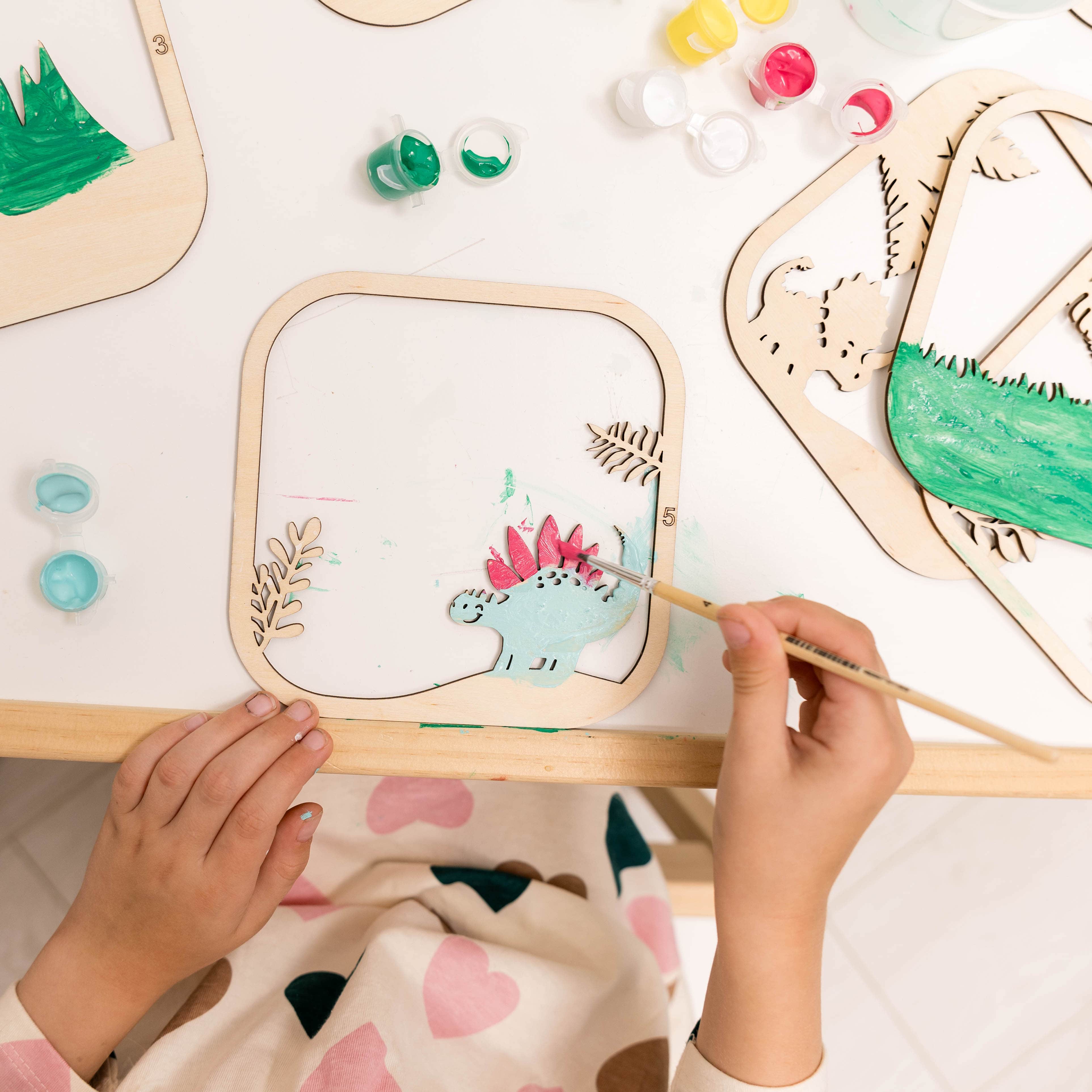 Child painting a wooden craft with a dinosaur design on a light surface.