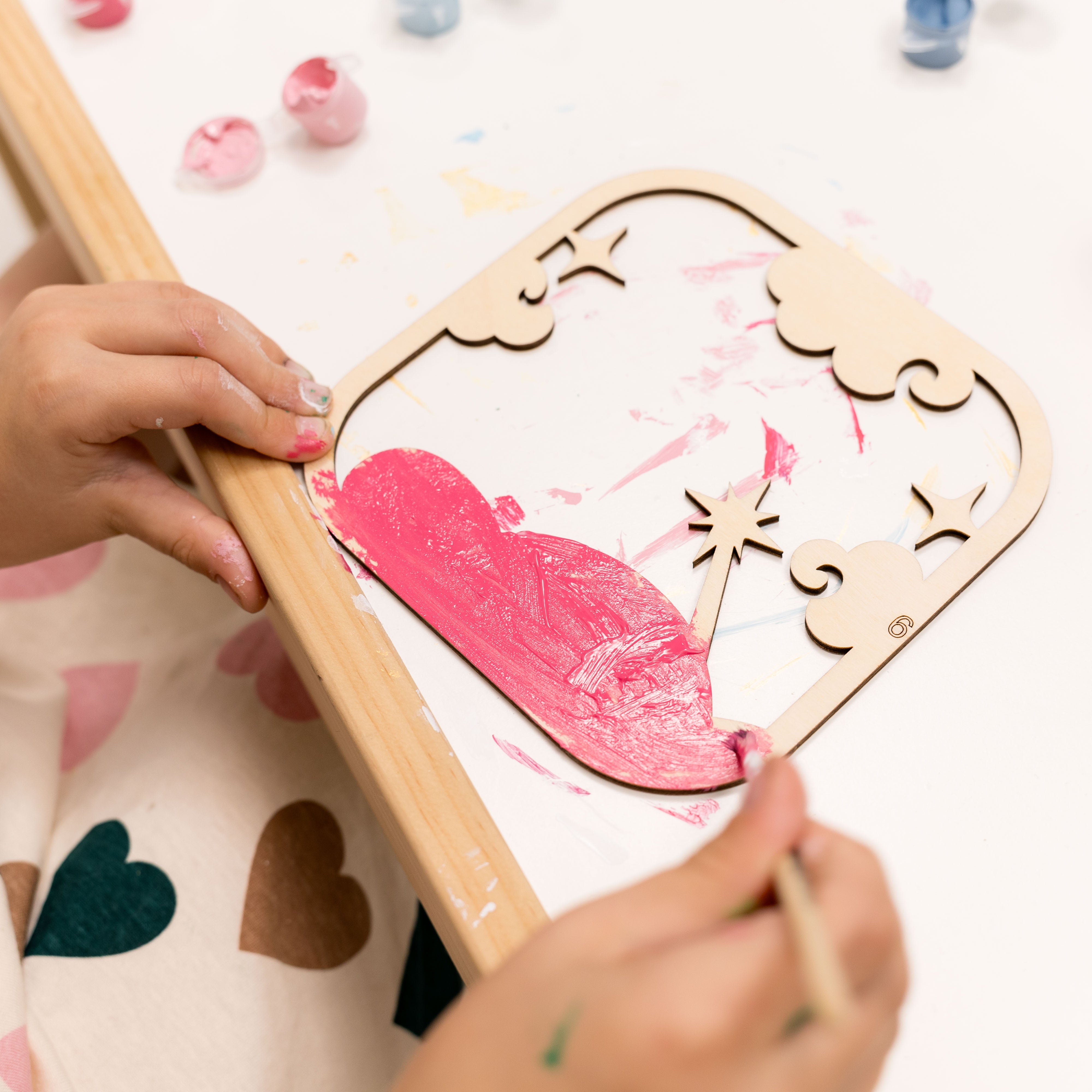 Child painting a pink heart on a wooden frame with colorful paint splashes.