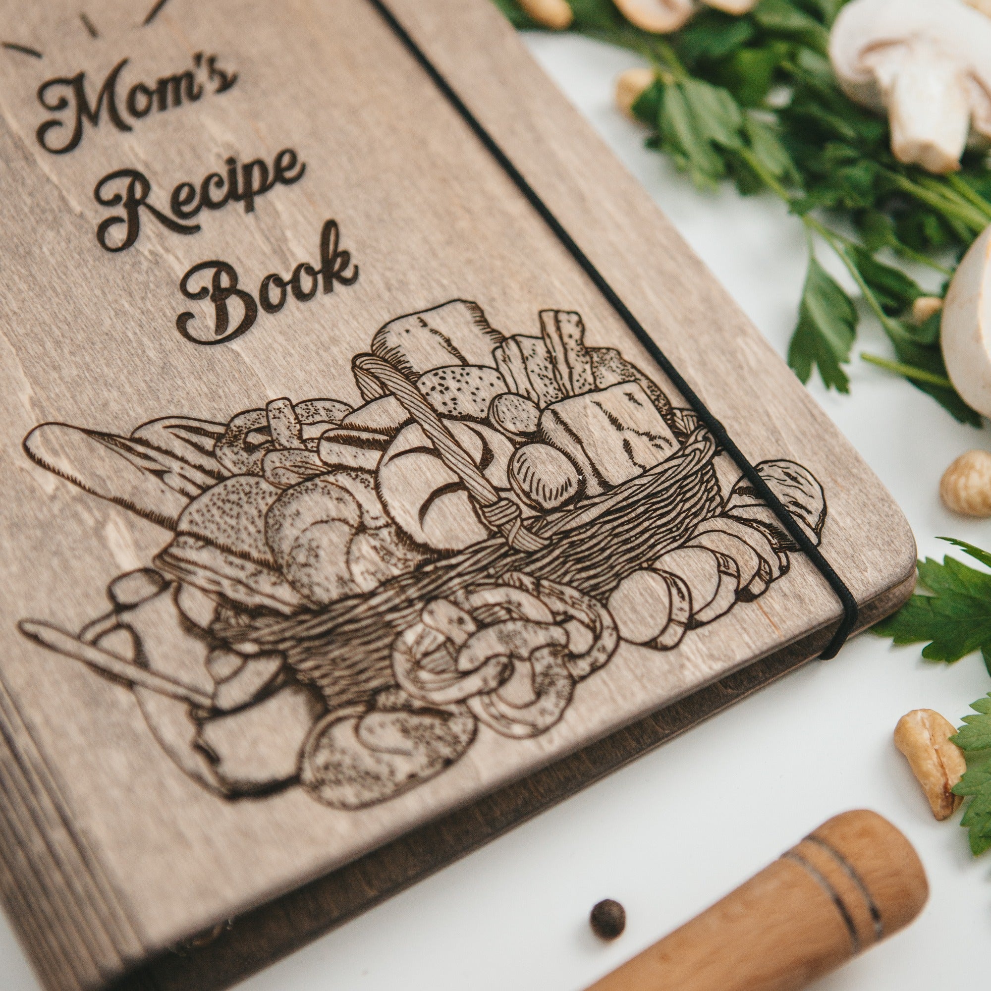 Wooden recipe book titled 'Mom's Recipe Book' with a engraved illustration of mushrooms on a white surface.