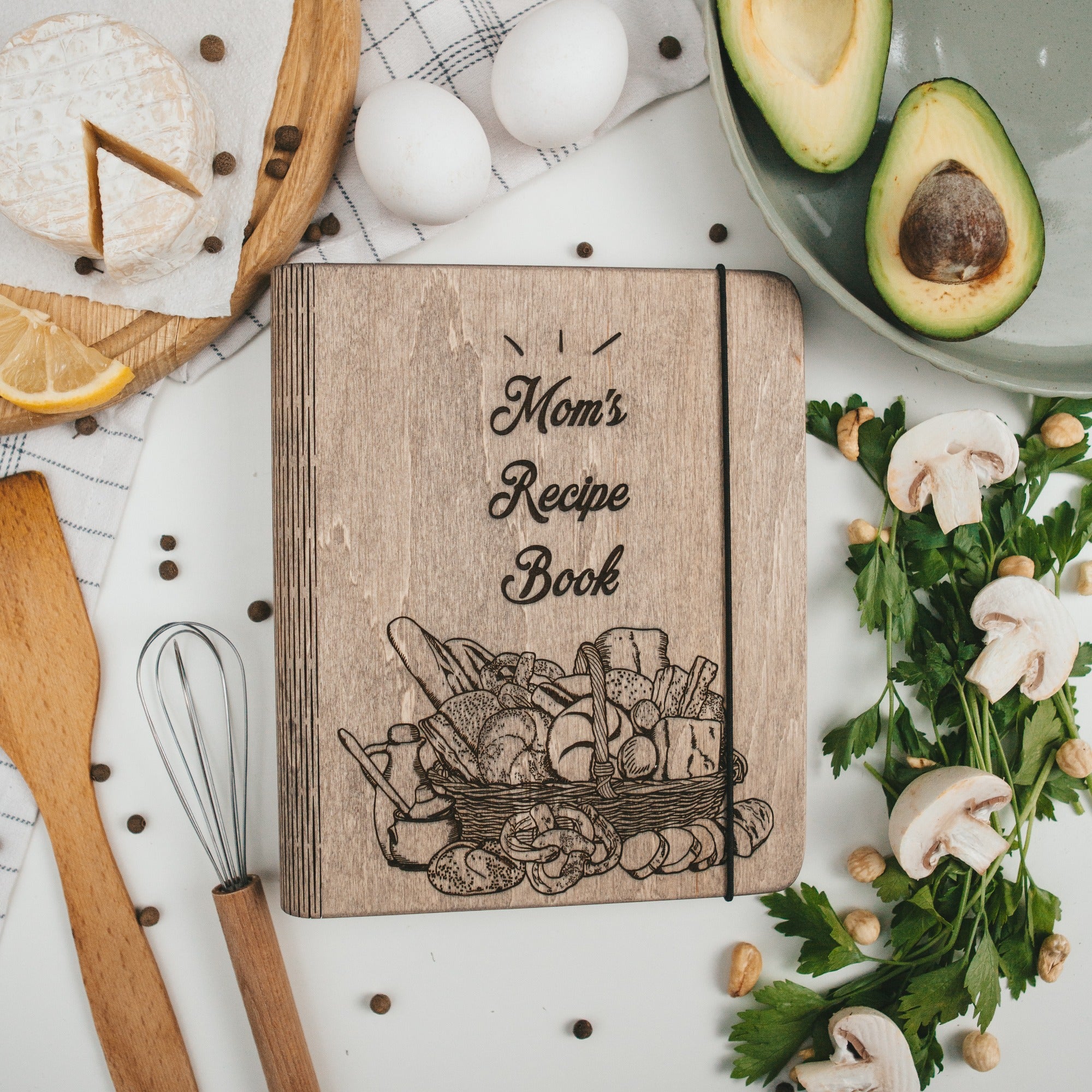 Wooden recipe book titled 'Mom's Recipe Book' surrounded by kitchen items on a white surface.