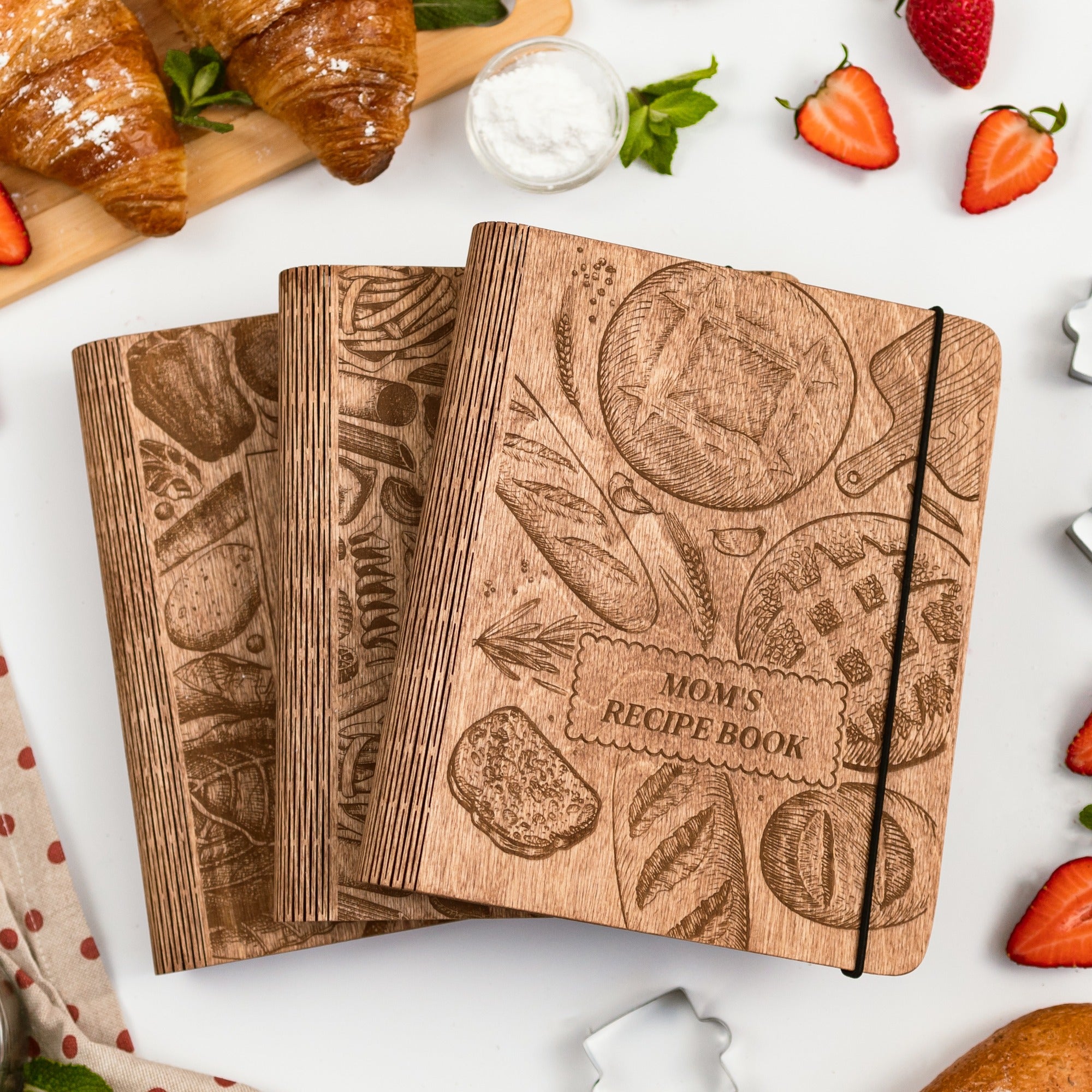 Wooden recipe book with bread design on a white surface with strawberries and croissants.