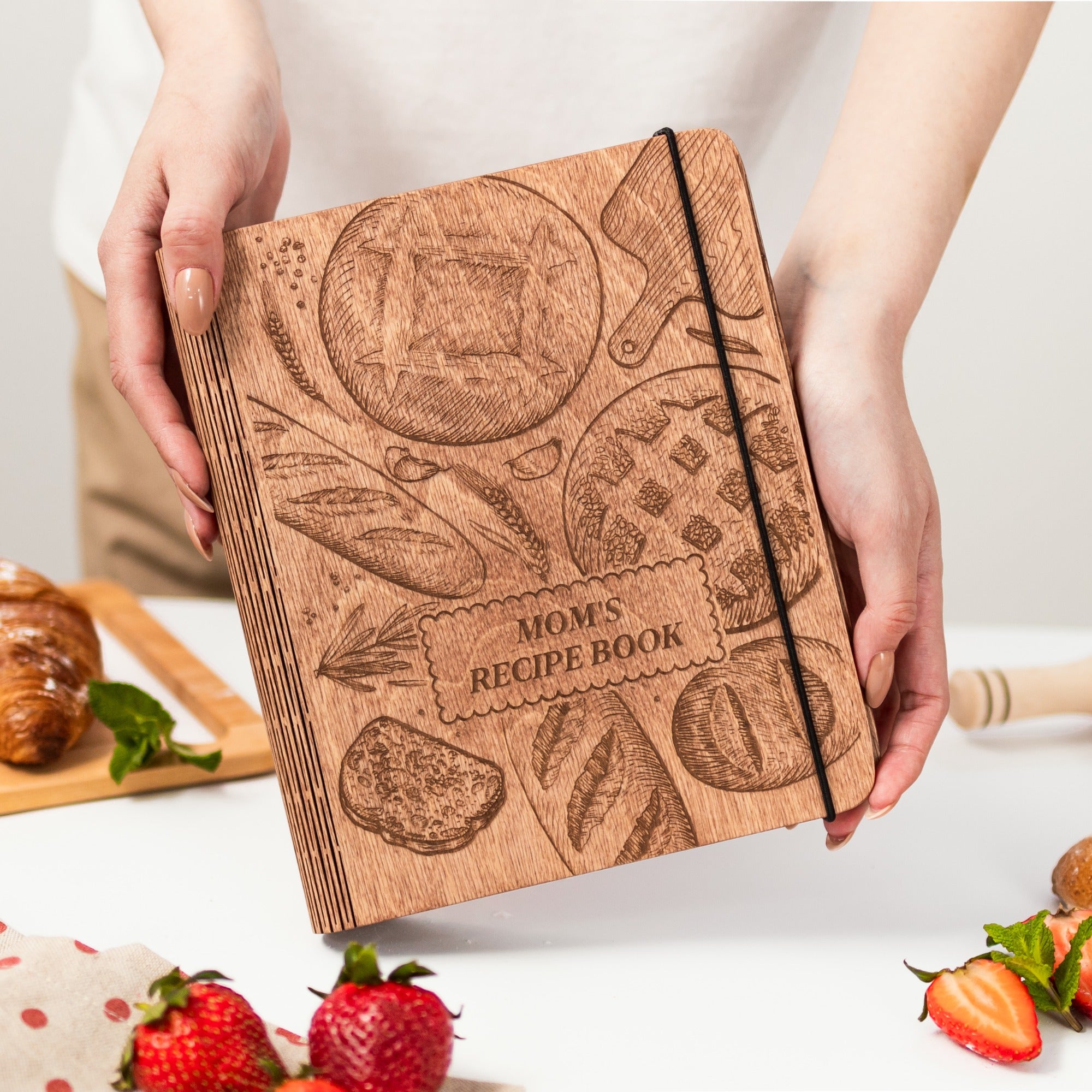 Wooden recipe book titled 'Mom's Recipe Book' held by hands with a kitchen setting in the background.