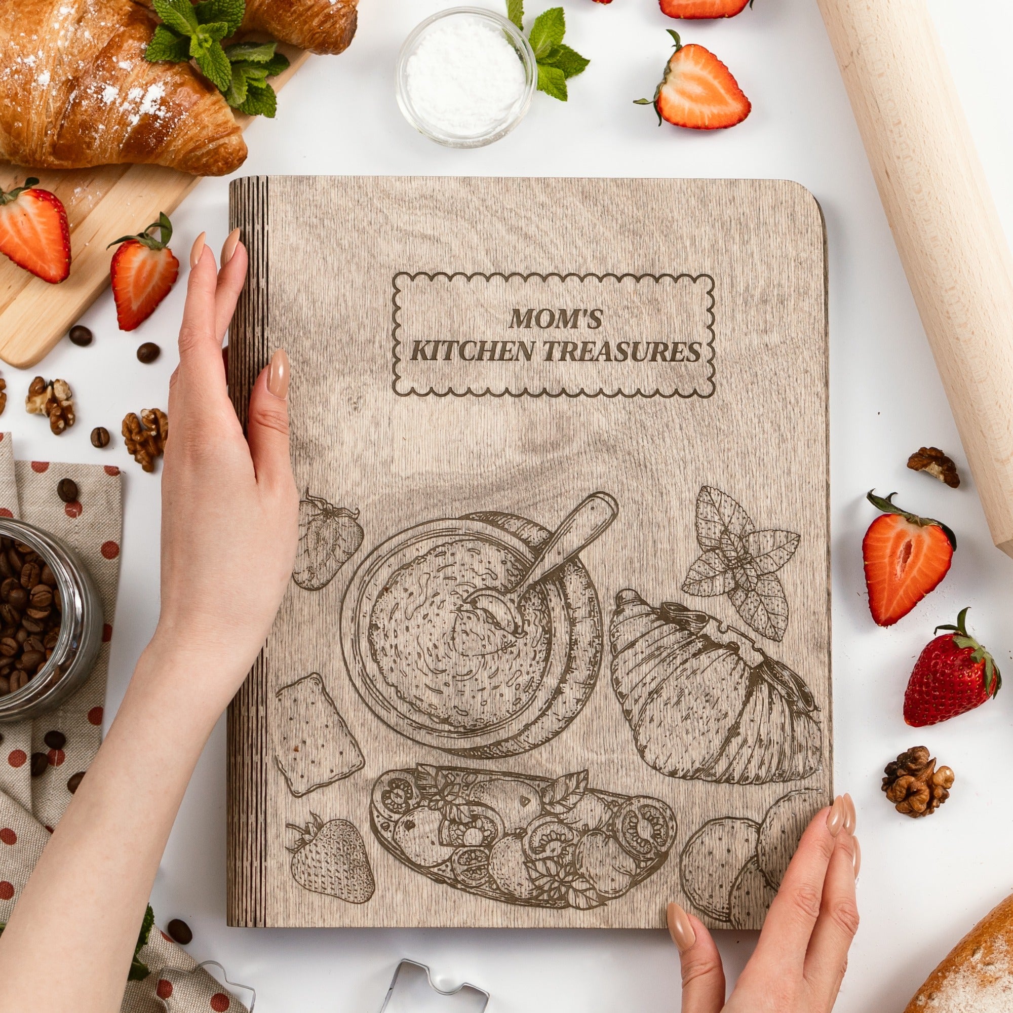 Cookbook titled 'Mom's Kitchen Treasures' held by hands on a kitchen counter with food items around.