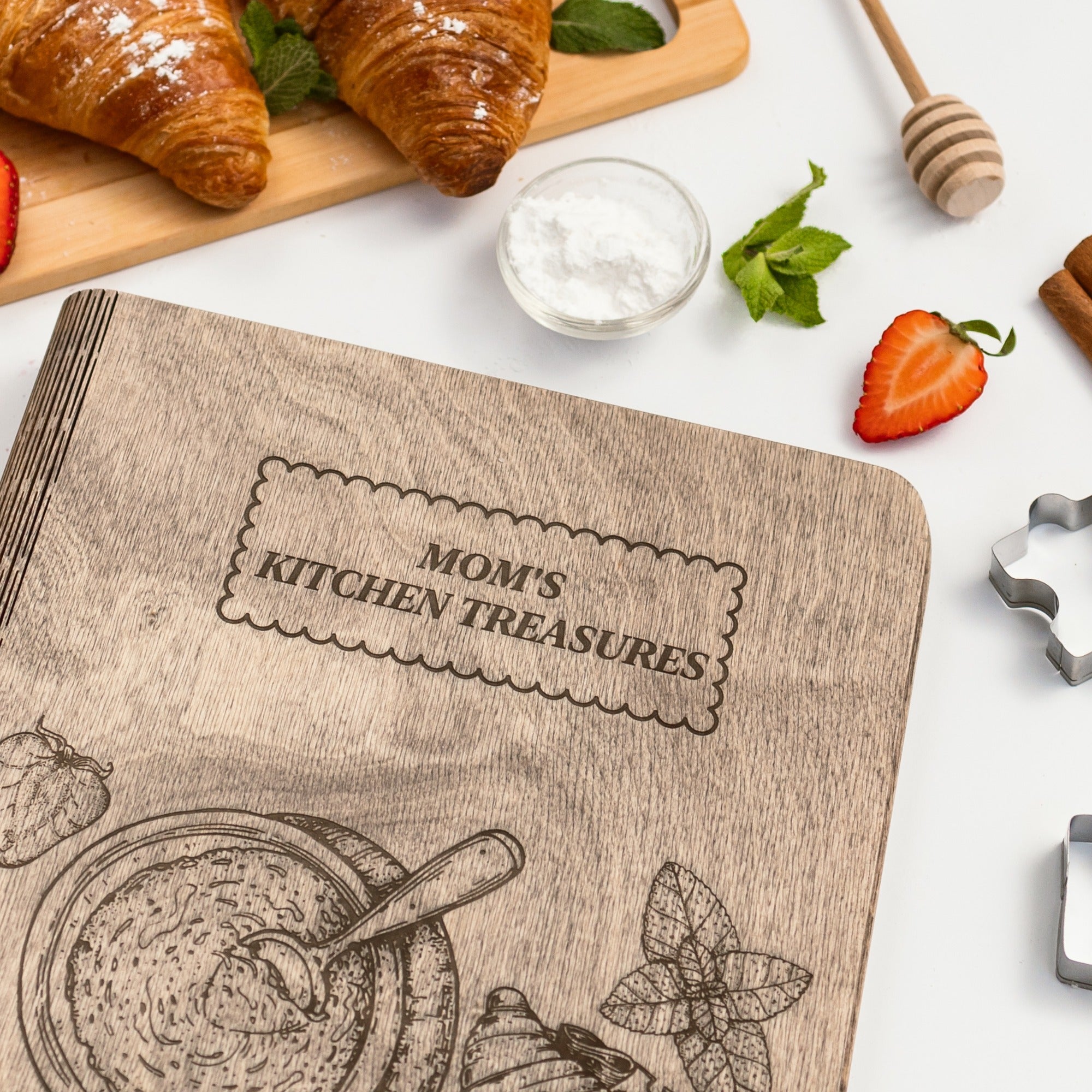 Cookbook titled 'Mom's Kitchen Treasures' with a wooden cover on a white background with food items.