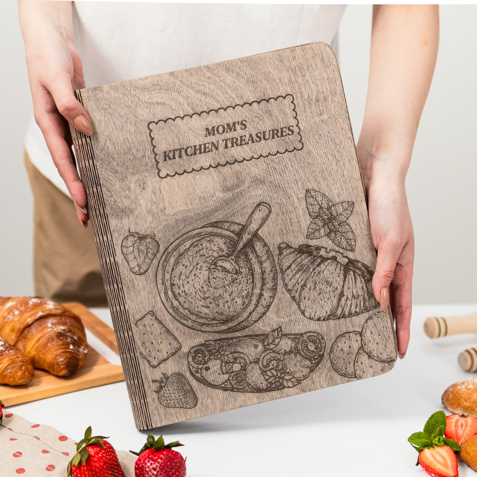 Cookbook titled 'Mom's Kitchen Treasures' held by a person with food items on a table.