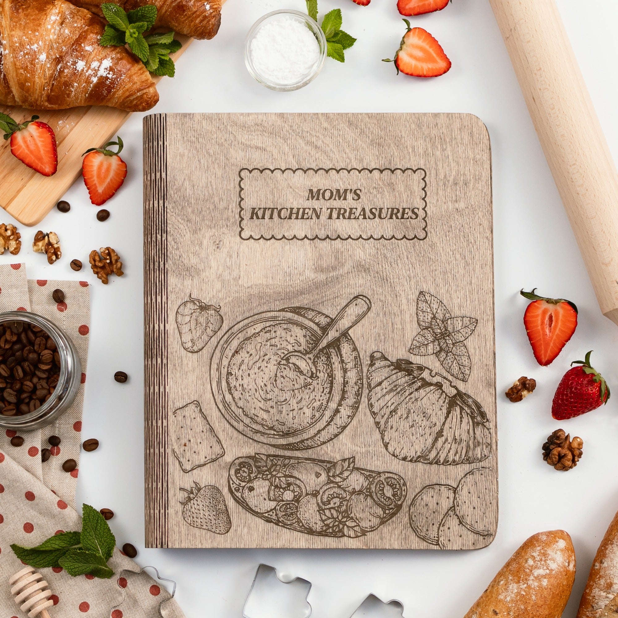 Cookbook titled 'Mom's Kitchen Treasures' surrounded by kitchen items on a white surface