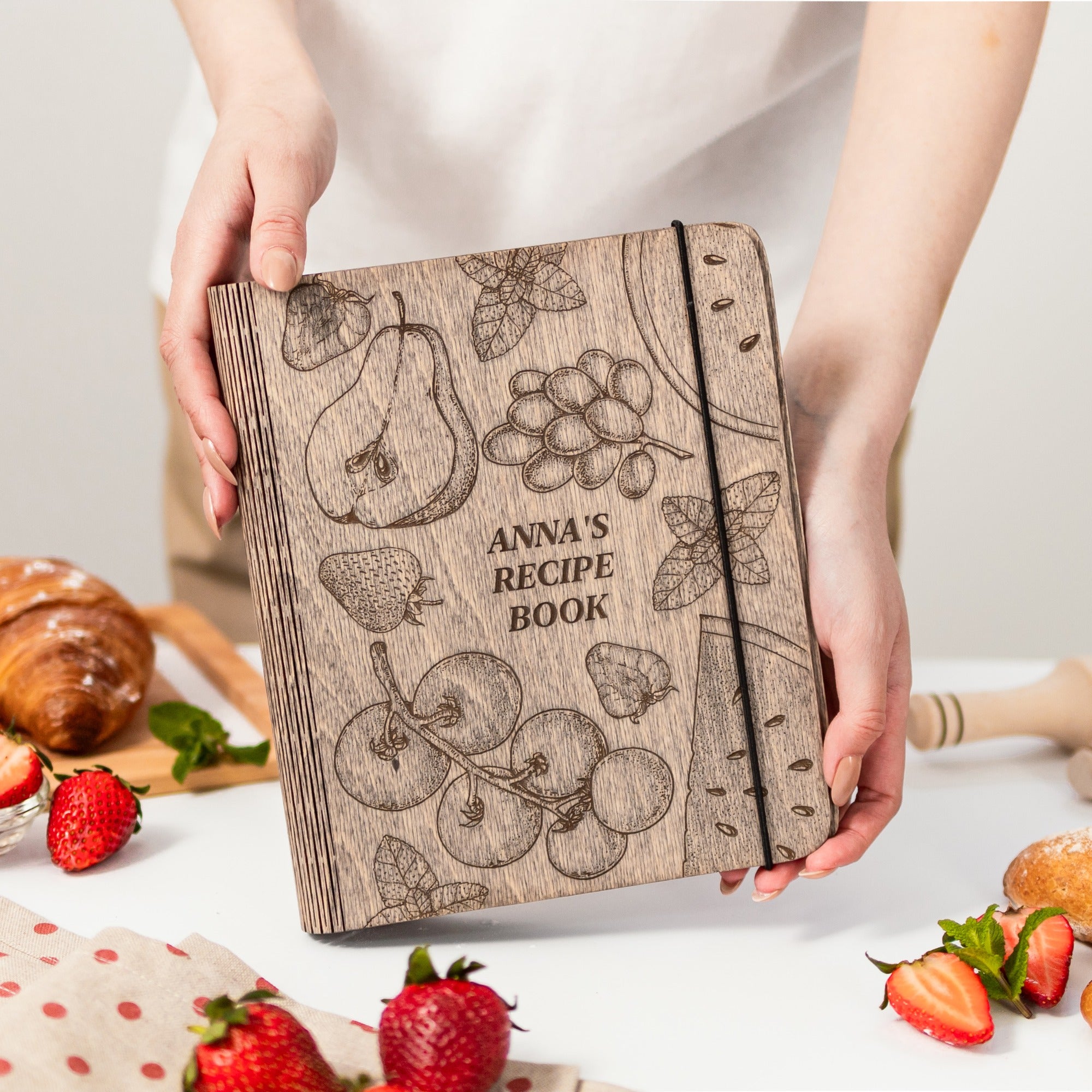 Wooden recipe book with fruit illustrations held by a person, surrounded by strawberries and pastries on a light background.